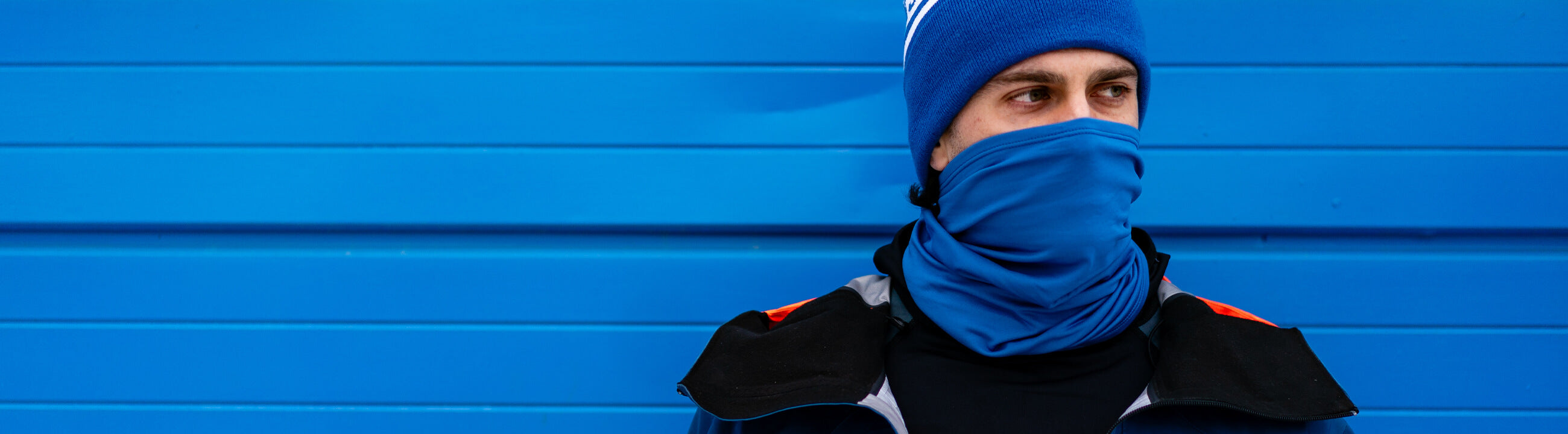Man wearing a Royal Blue Neck Gaiter posted agaist a Blue Garage door.