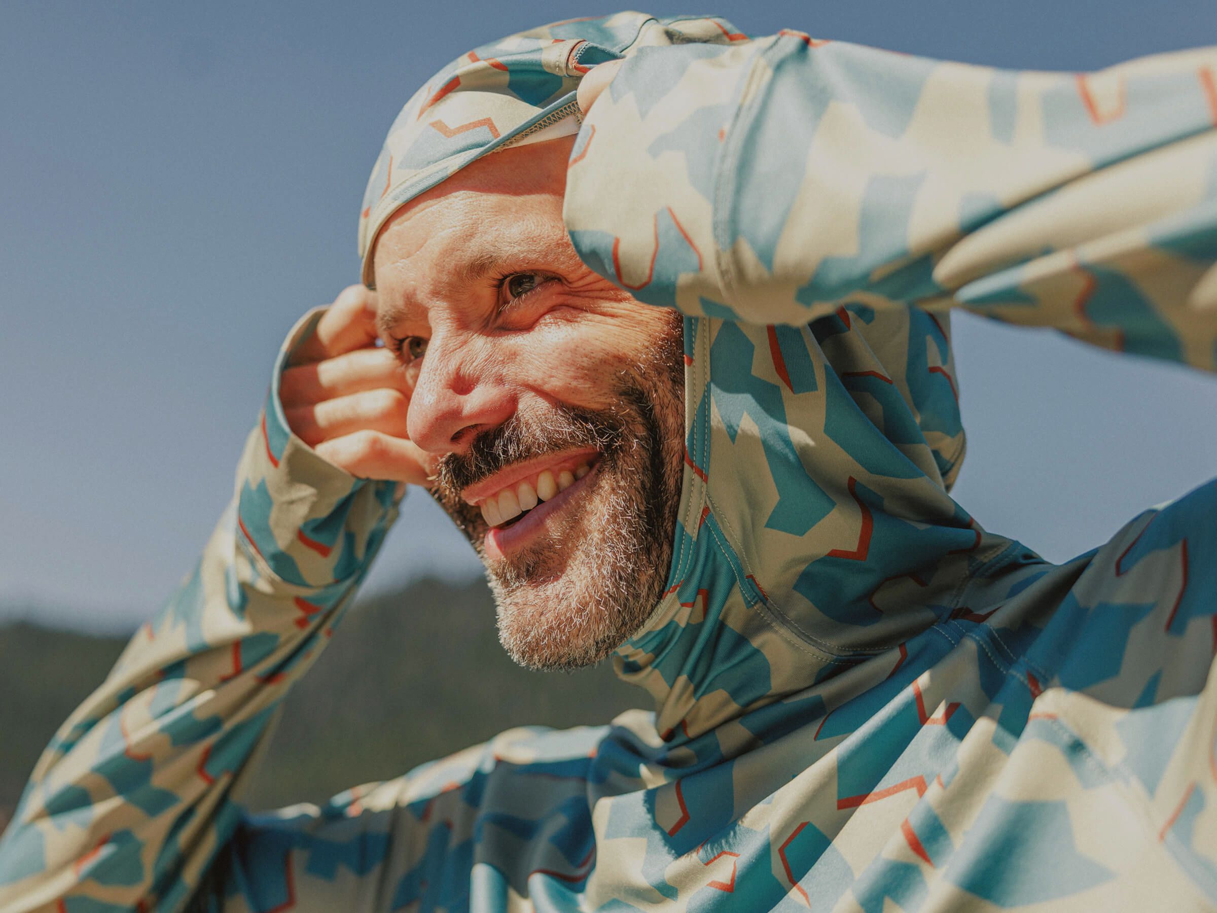 Man wearing a BlackStrap Brackish Sun Hoodie in Dimentical Sagebrush colorway with the hood on and mountains in the background.