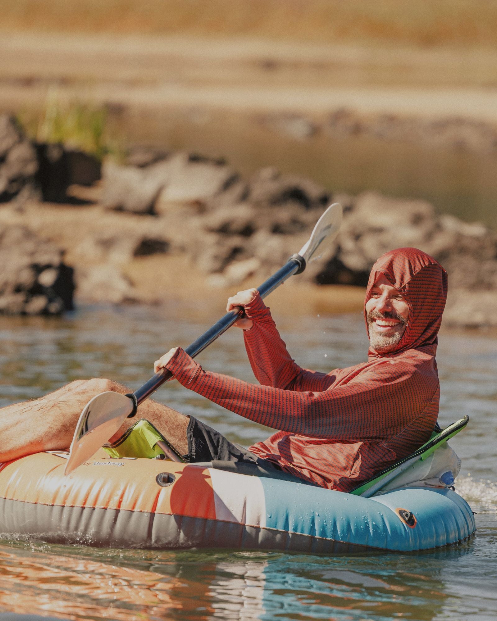 Person in the BlackStrap Brackish Sun Hoodie paddling a kayak on a calm body of water with rocks in the background.
