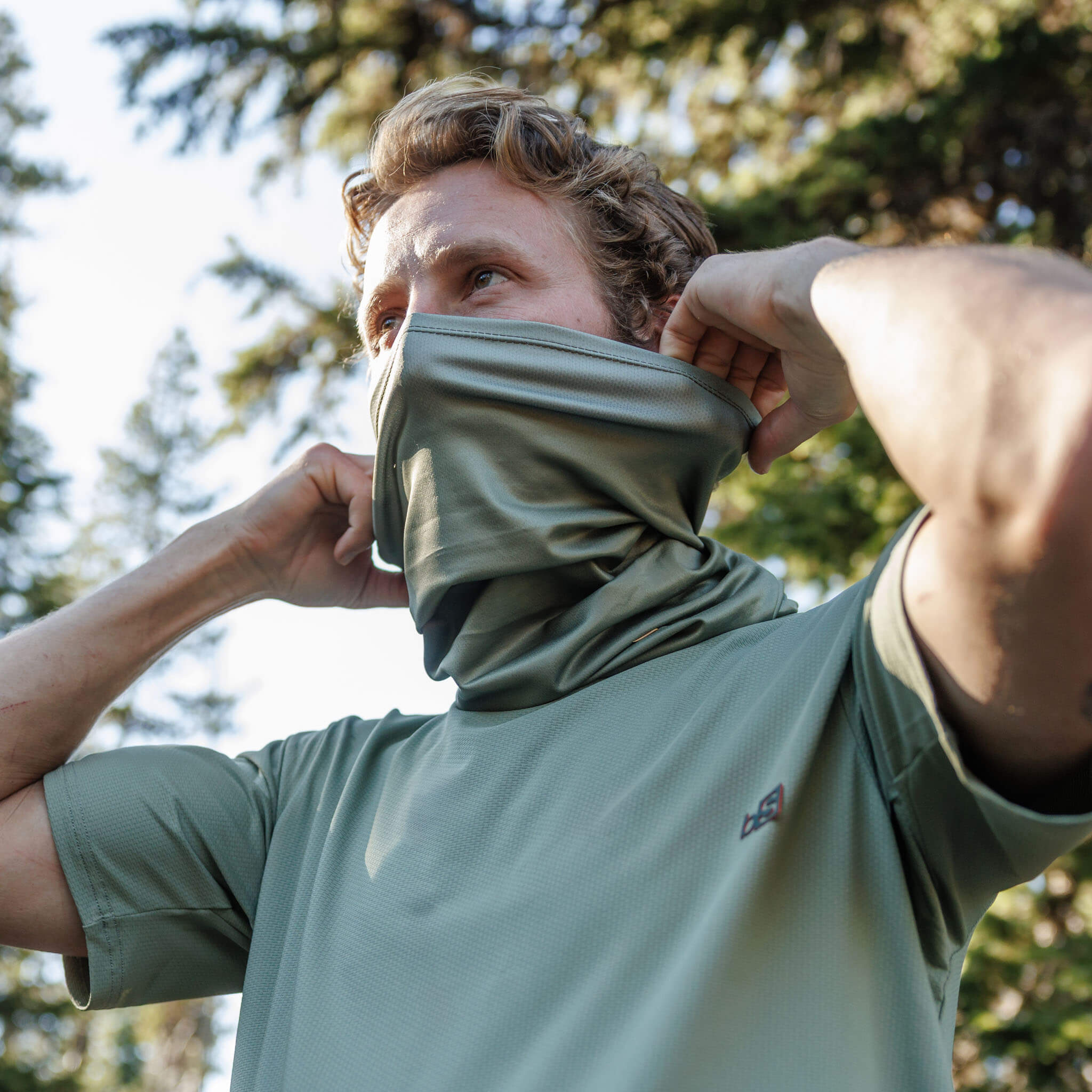 Man pulling a BlackStrap Daily Tube neck gaiter up over his face in sage green, outdoors among pine trees