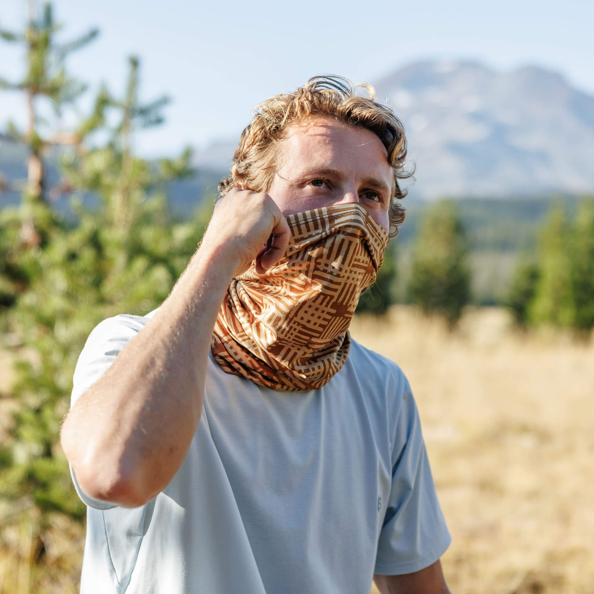 Man pulling a BlackStrap Daily Tube neck gaiter up over his face, with a mountain peak in the background #color_scribble lark
