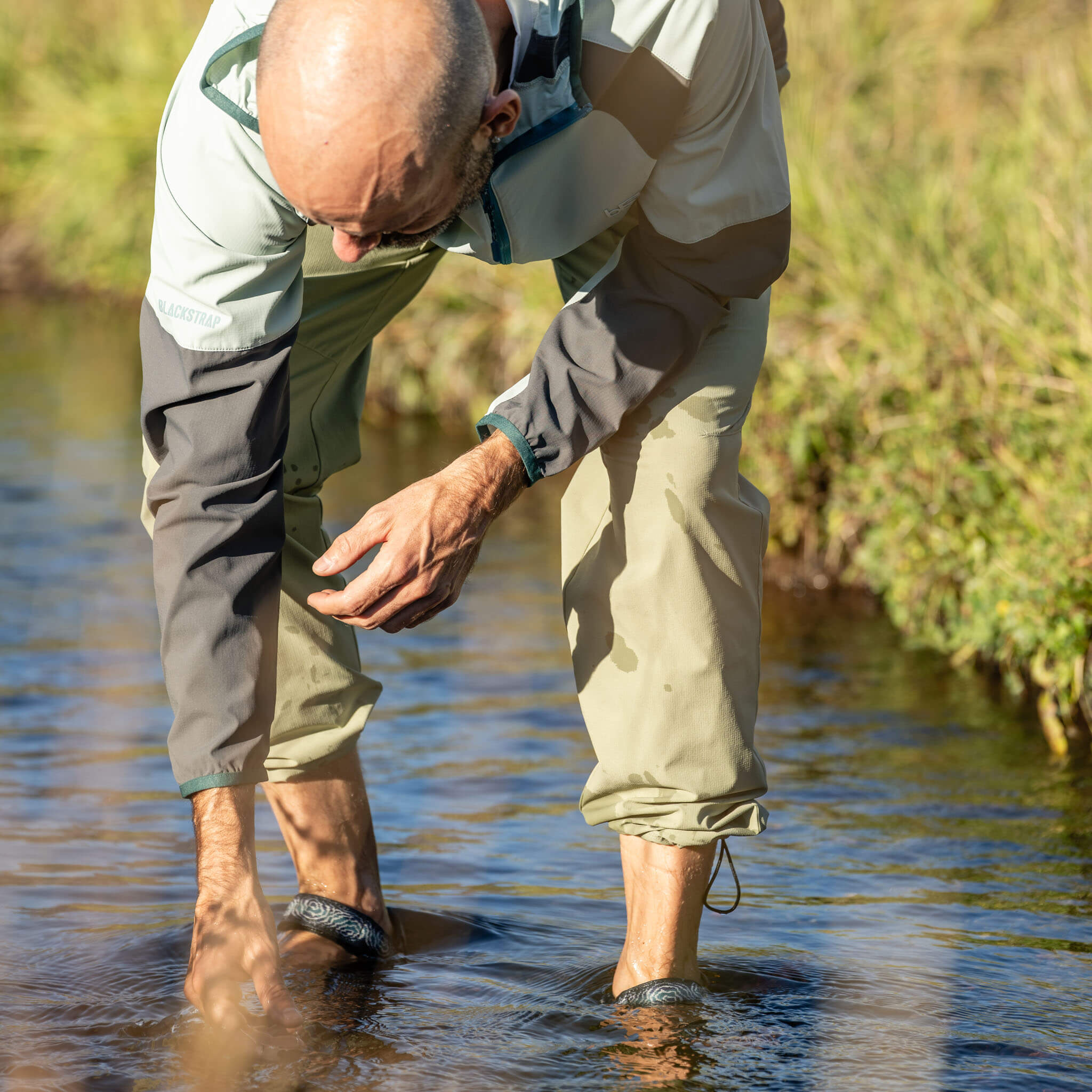 Man leaning over to touch the water while wading in a stream wearing BlackStrap approach pants rolled up at the ankles and sport sandals, with a Cirq full zip wind jacket and grassy banks behind him. #scolor_spruce