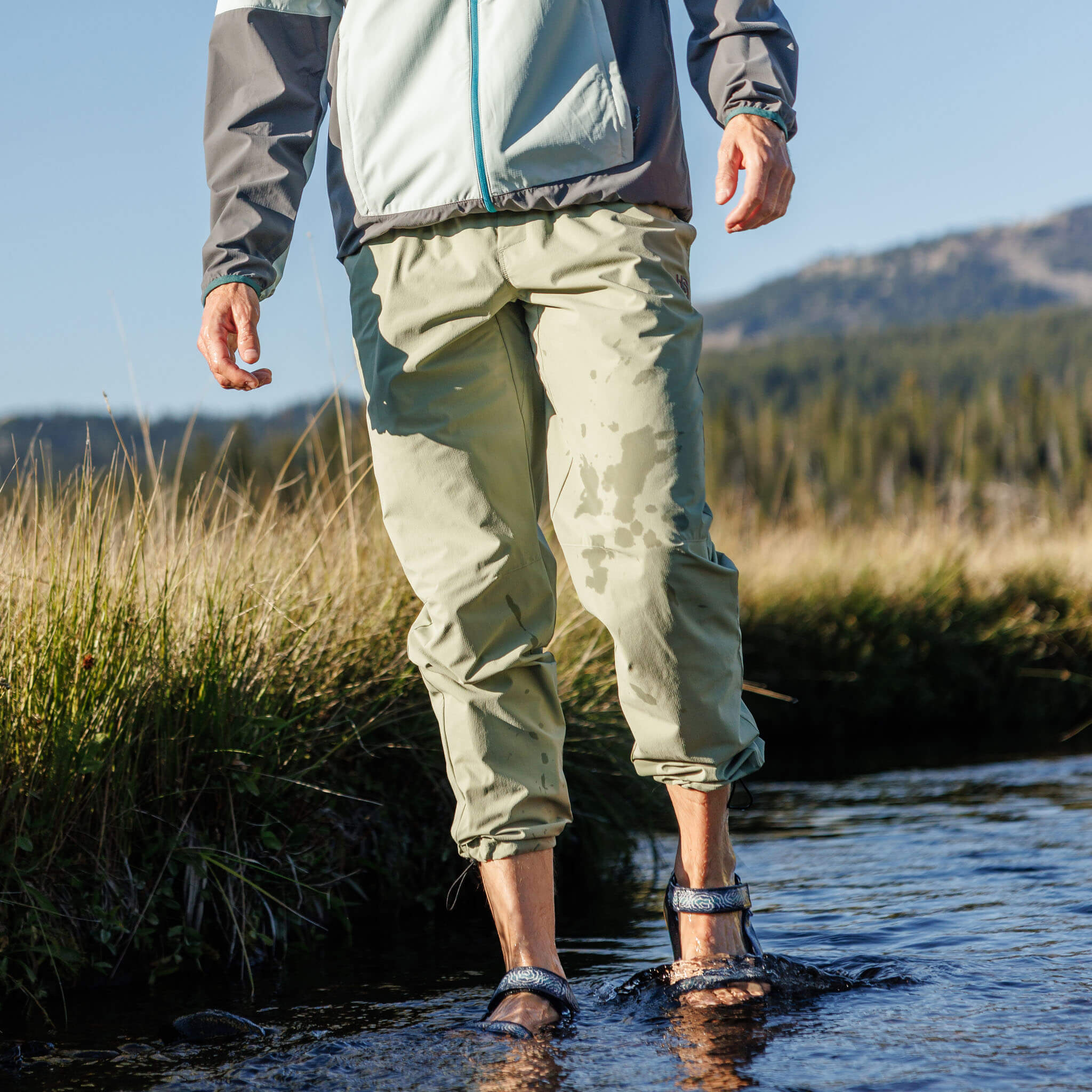 Man wading through a stream wearing BlackStrap approach pants rolled up at the ankles and sport sandals, with a wind jacket on top, tall grasses and mountains in the background. #color_sagebrush