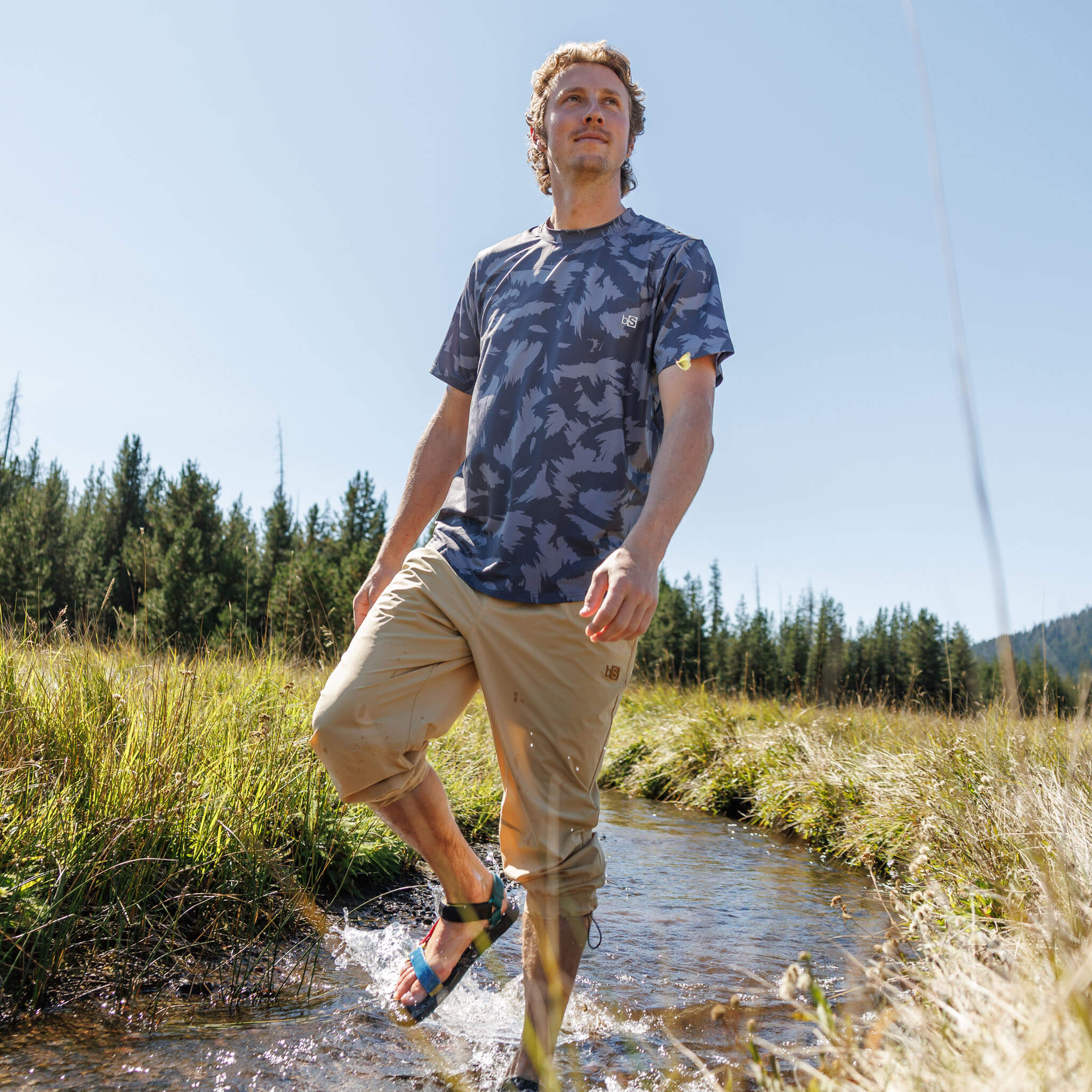 Man walking through a shallow stream wearing BlackStrap approach pants rolled up at the knees and sport sandals, paired with a Slashed Smoldered Voyage t-shirt, with pine trees and open sky behind him. #color_lark