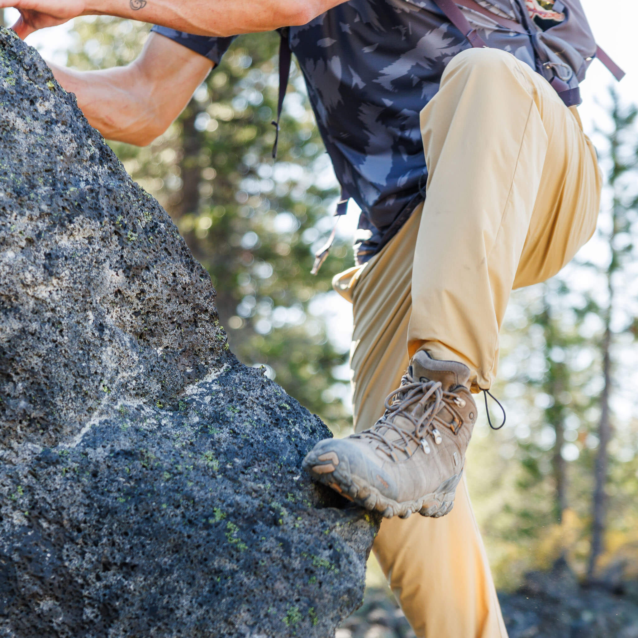 Close-up of a man scrambling up a boulder wearing BlackStrap approach pants and hiking boots, with a slash Smoldered voyage trail shirt and backpack visible above. #color_lark