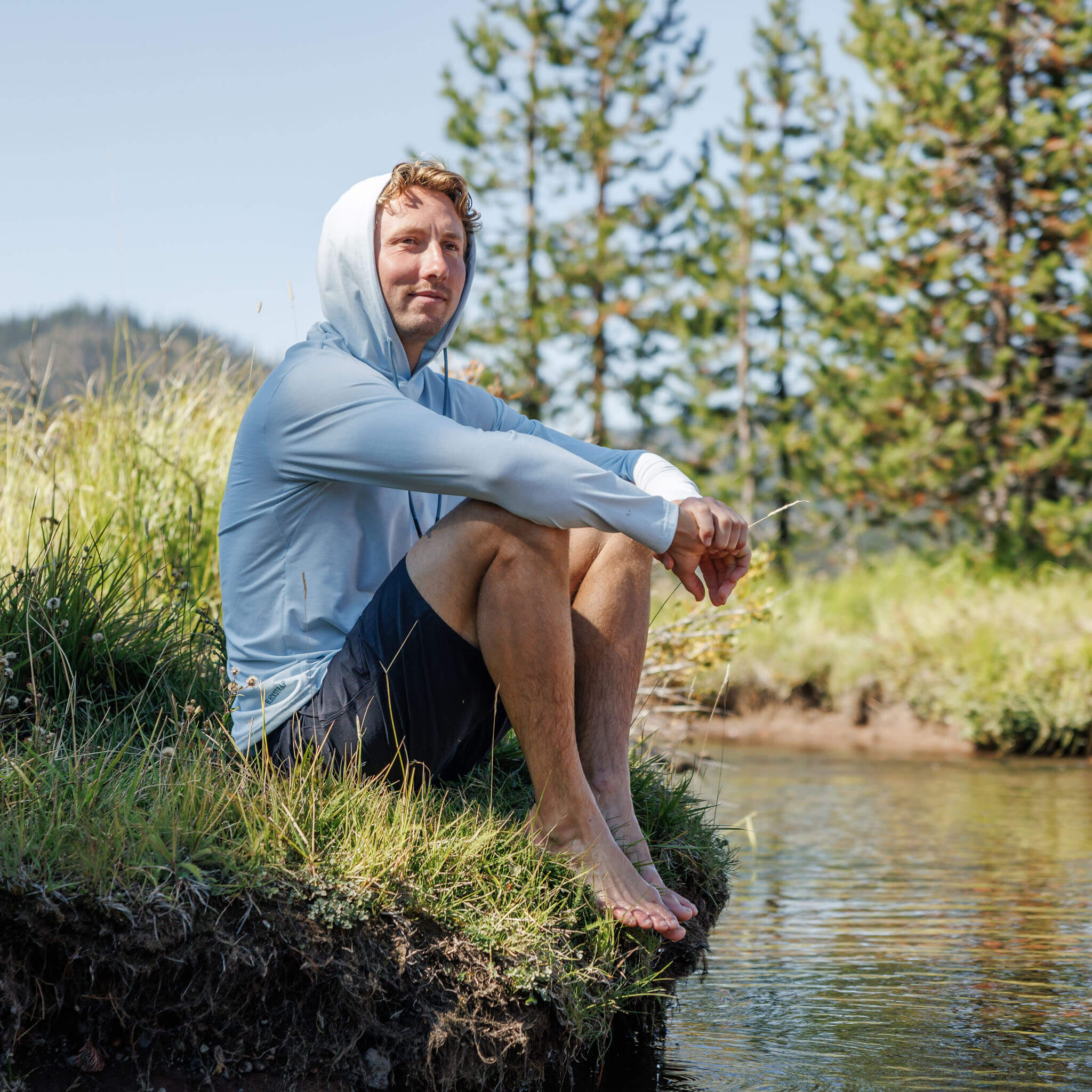 Man sitting on a grassy riverbank with hood up wearing the BlackStrap Men's Aspect Hoodie Long Sleeve #color_stratus