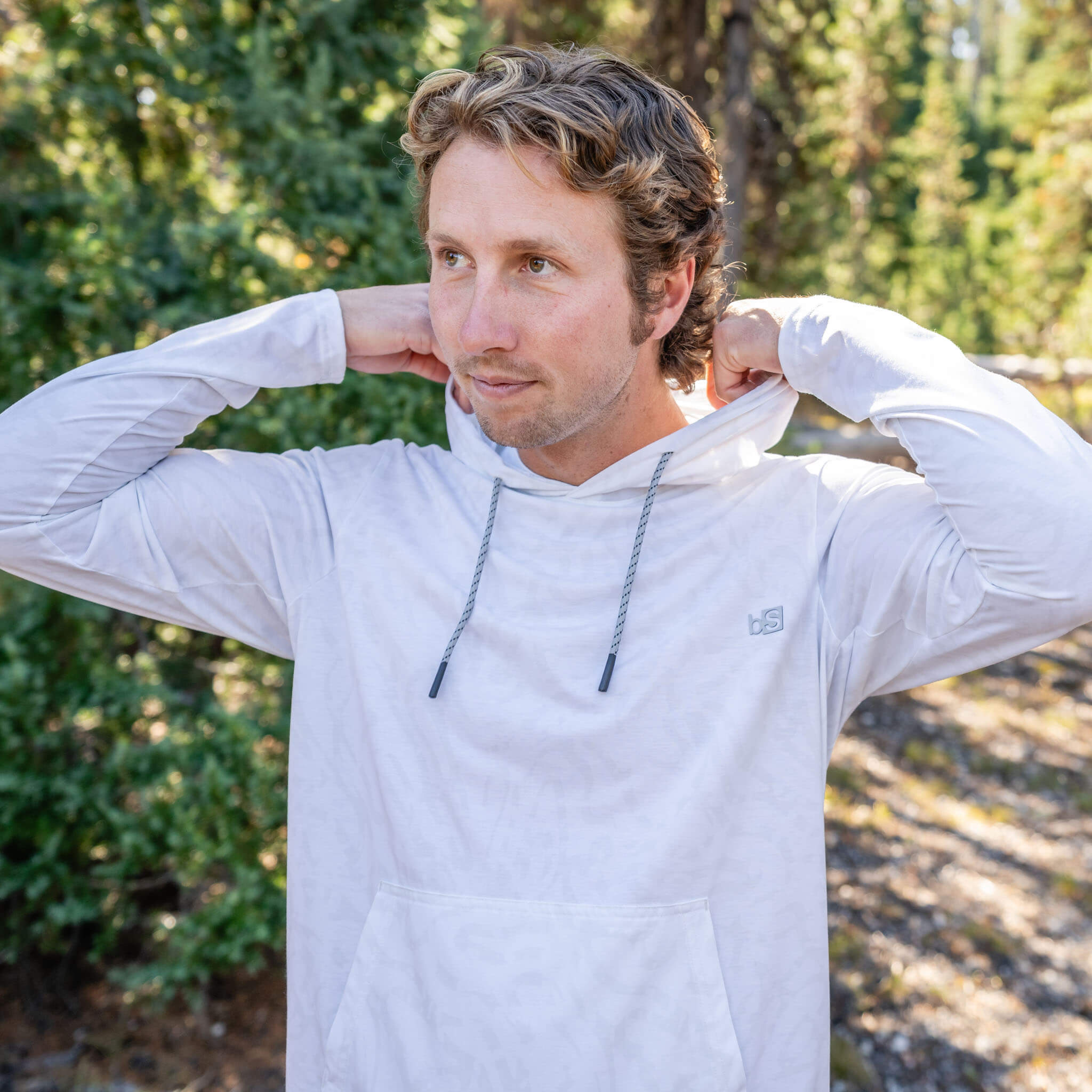 Close-up of a man standing in a pine forest, adjusting the hood of the BlackStrap Men's Aspect Hoodie Long Sleeve #color_ripple ash