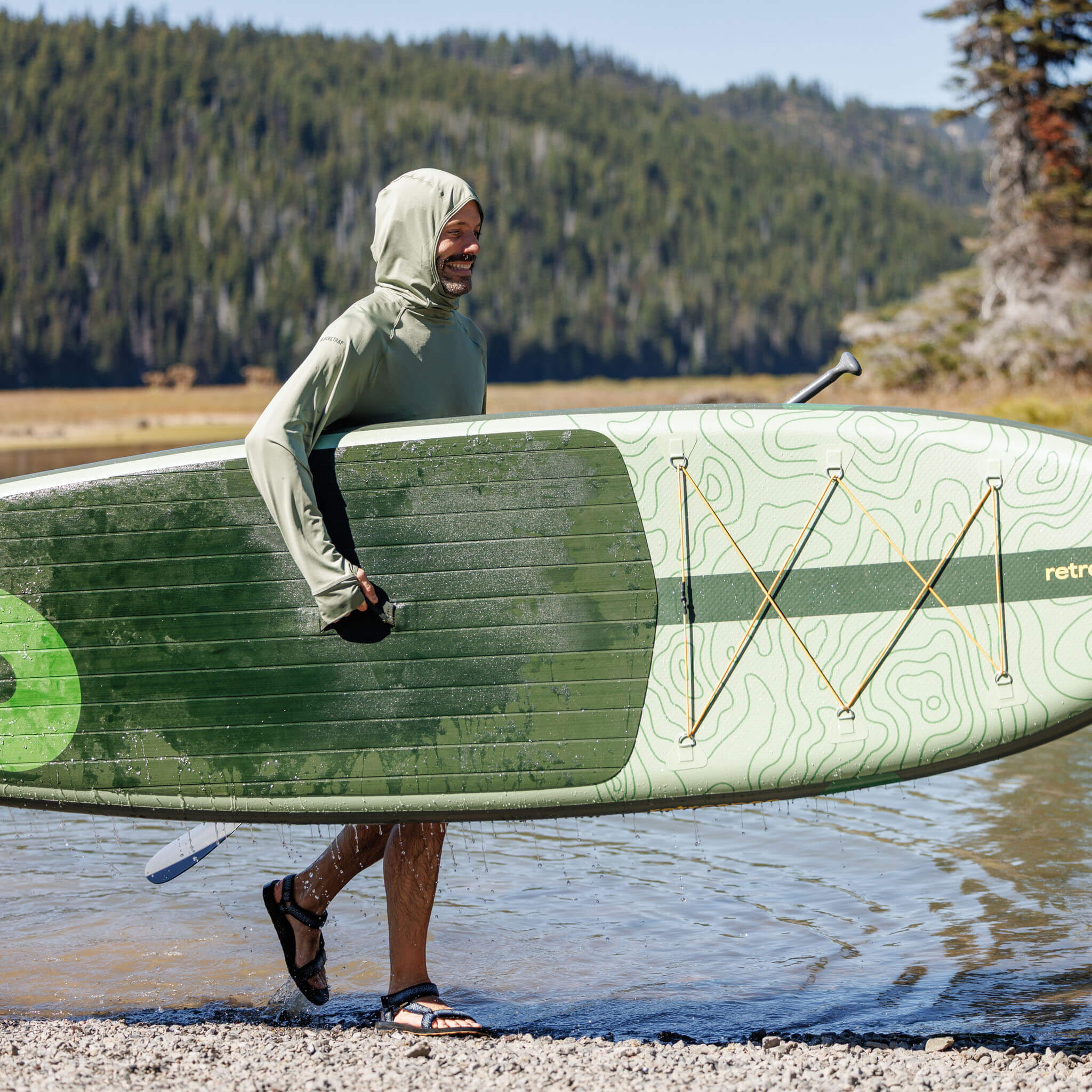 Person wearing the BlackStrap Brackish Sun Hoodie and carrying a green paddleboard with a forest and lakeshore in the background