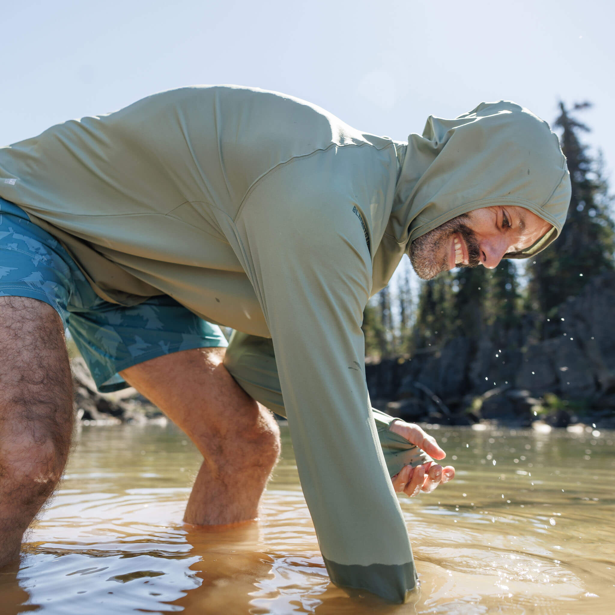 Man reaching into a shallow mountain lake wearing the BlackStrap Men's Brackish Long Sleeve Hoodie with hood up #color_sagebrush