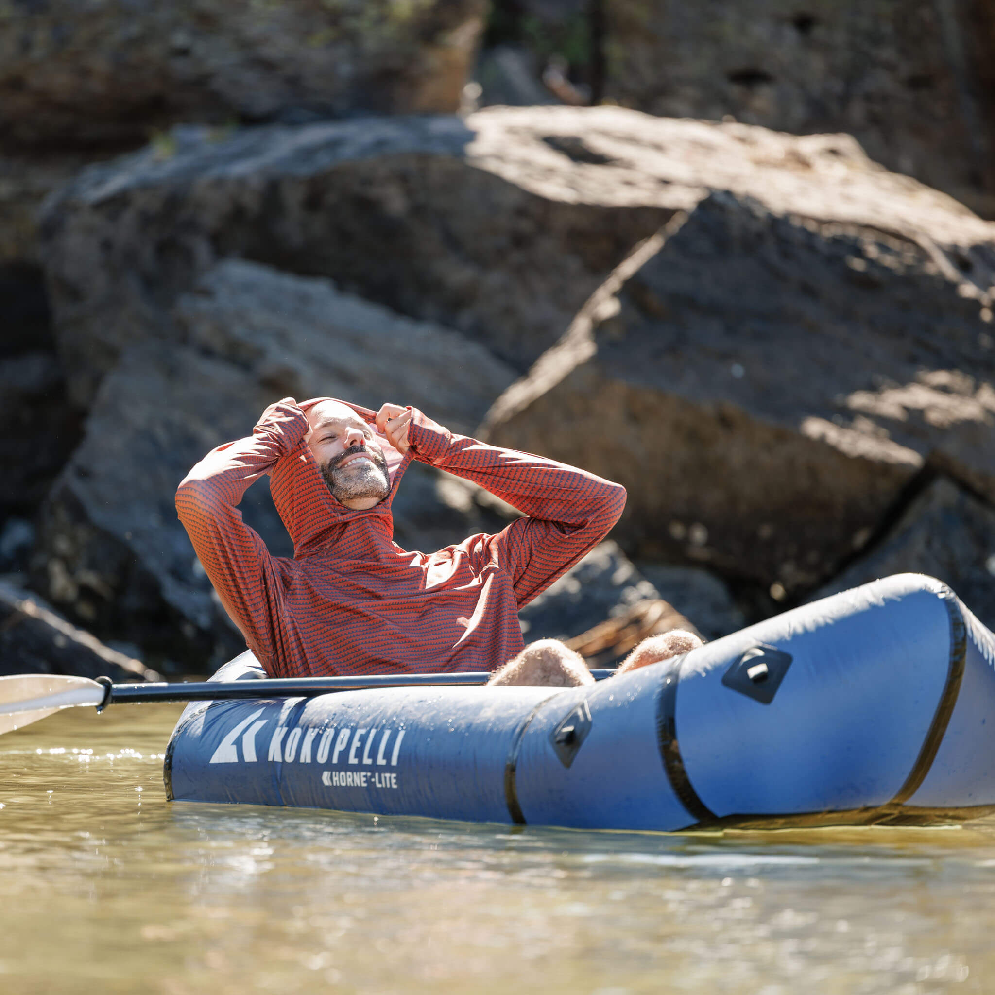 Person wearing the BlackStrap Brackish sun Hoodie in a blue kayak with 'Kokopelli' branding, surrounded by rocks and water. #color_sharktooth jory