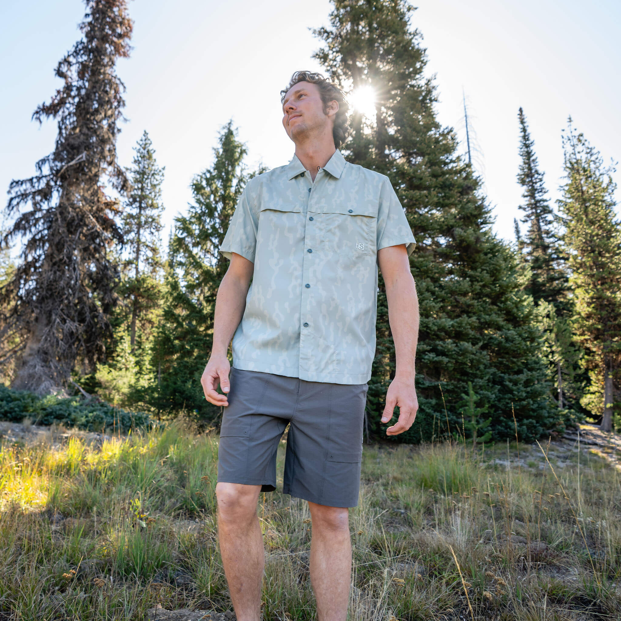 Man standing in an alpine meadow looking upward, wearing a Corally Spruce short-sleeve BlackStrap camp shirt with chest pockets and utility shorts, with pine trees and sunlight behind him.