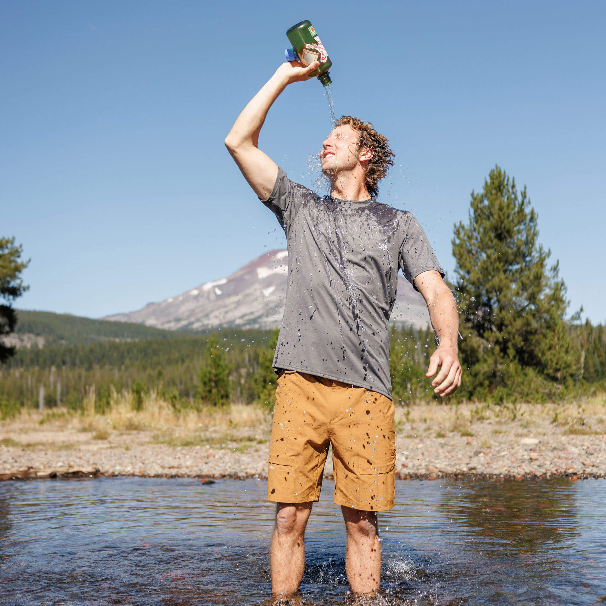 Man standing in a shallow river pouring water over his head from a bottle, wearing BlackStrap utility shorts and a Voyage t-shirt, with pine trees and a mountain in the background.