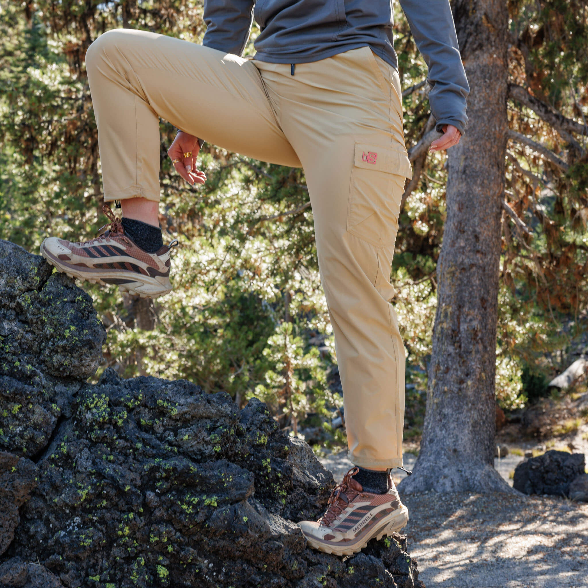 Woman stepping over rocks in a pine forest wearing BlackStrap approach pants with a cargo pocket and hiking shoes, with a long-sleeve layer on top. #color_lark