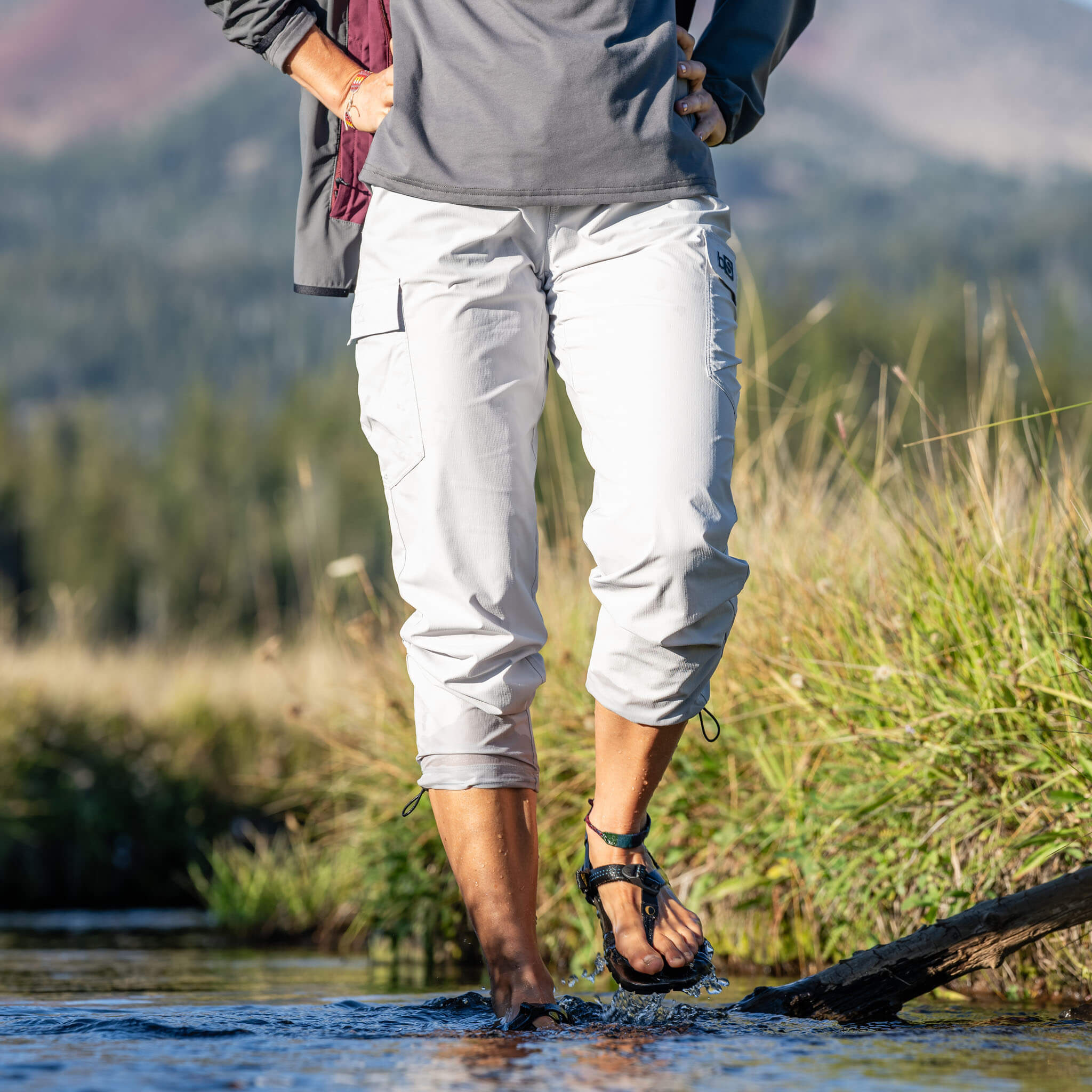 Woman wading through a shallow stream wearing BlackStrap approach pants rolled up to capri length and sport sandals, with mountains and tall grasses in the background #color_ash