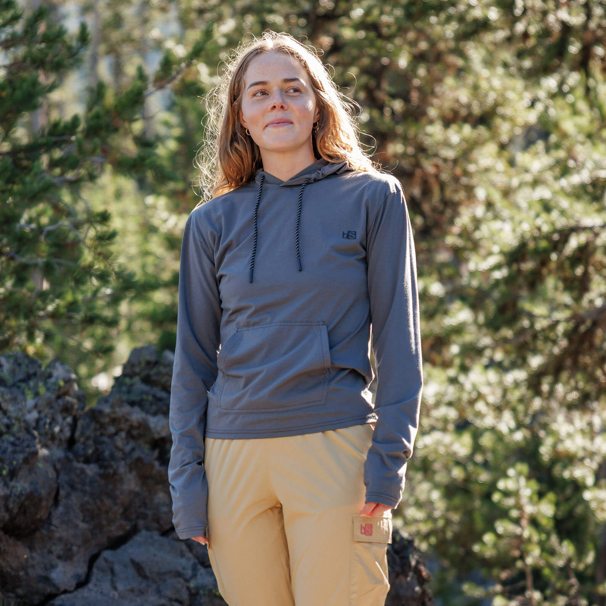 Woman standing among rocky alpine terrain wearing the BlackStrap Women's Aspect Hoodie Long Sleeve #color_smoldered