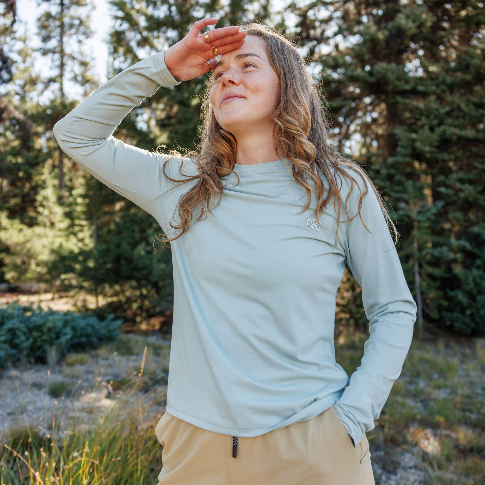 Woman shading her eyes from the sun in a pine forest wearing the BlackStrap Women's Aspect Long Sleeve #color_spruce