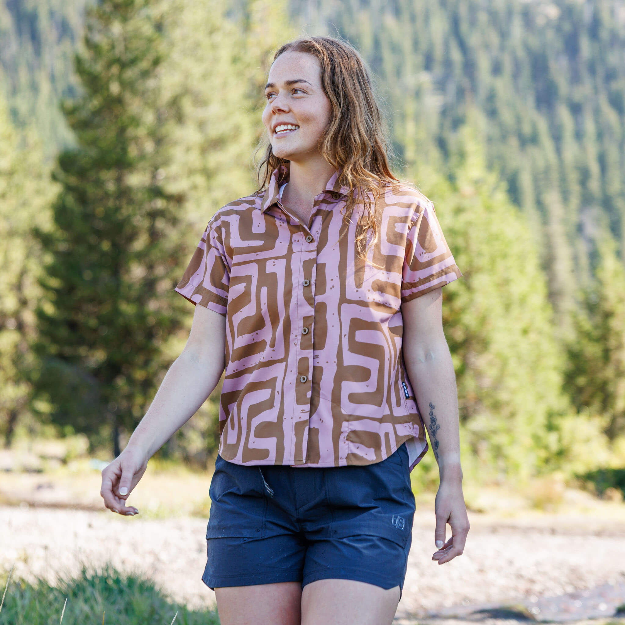 "Woman smiling and walking outdoors wearing a Blaize Canyon short-sleeve BlackStrap camp shirt with utility shorts, with pine trees and mountains in the background #color_blaize canyon