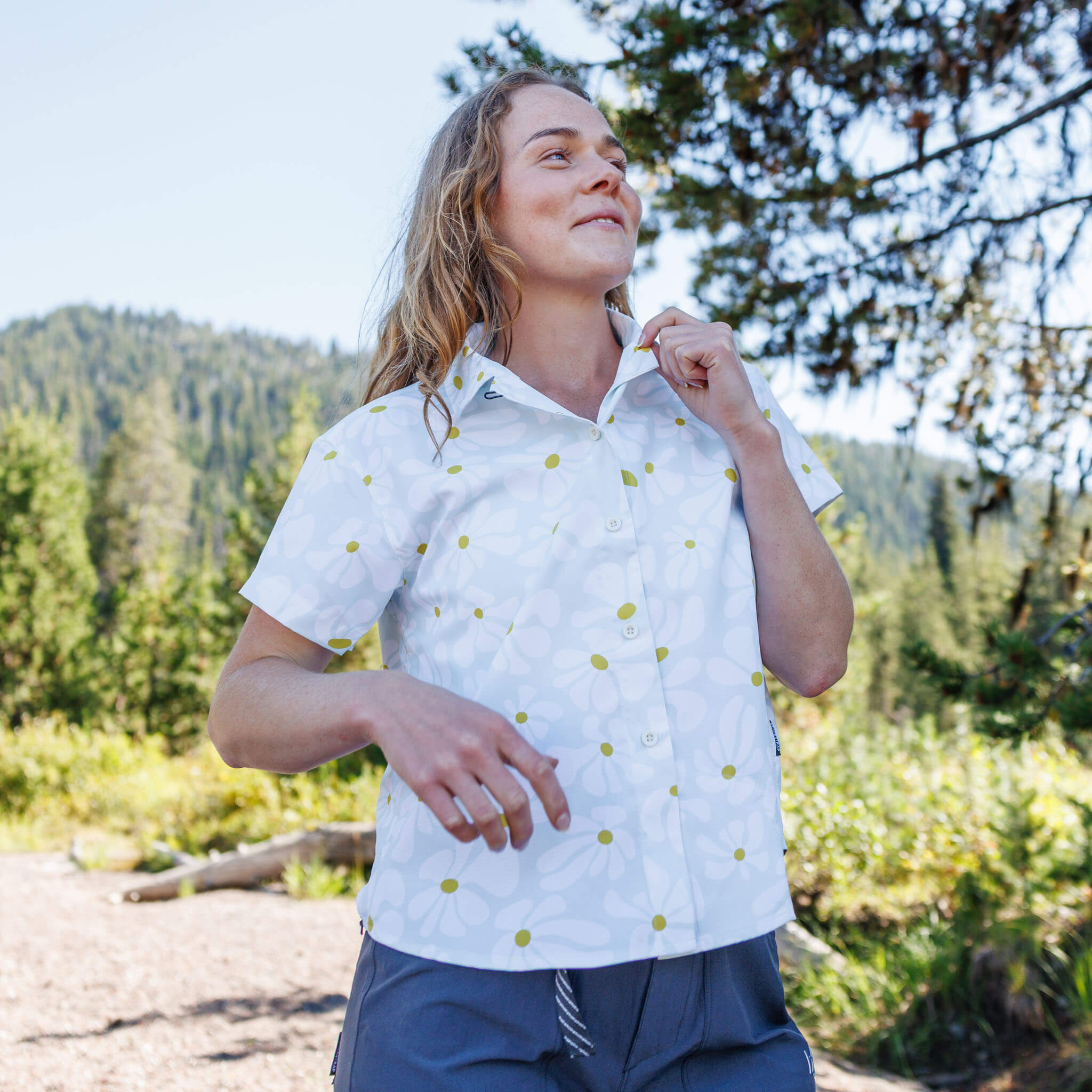 Woman on a forest trail adjusting the collar of a Boho Stratus short-sleeve BlackStrap camp shirt, with pine trees and mountains visible behind her. #color_boho stratus