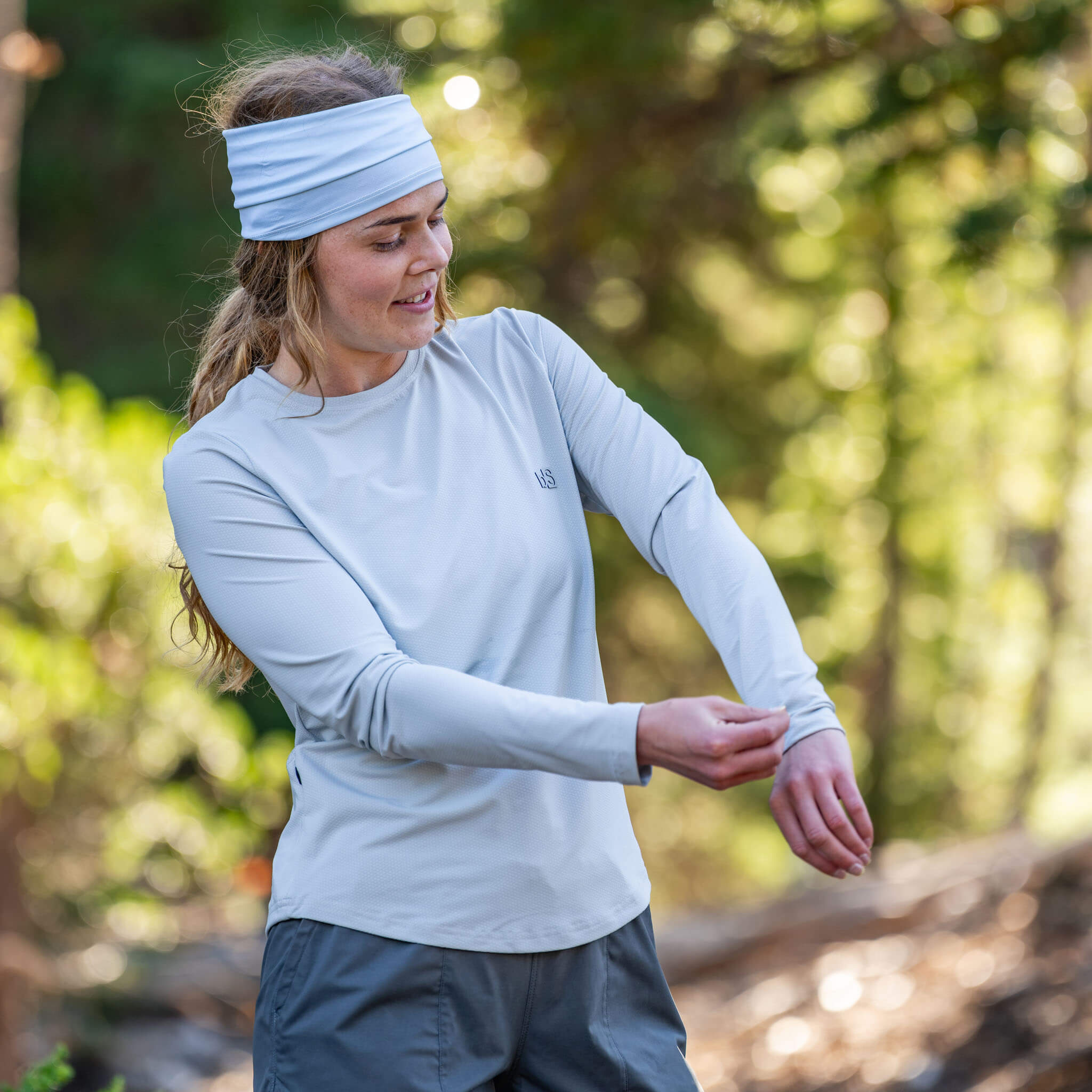 Woman adjusting her sleeve on a forest trail wearing the BlackStrap Women's Voyage Crew Long Sleeve #color_ash