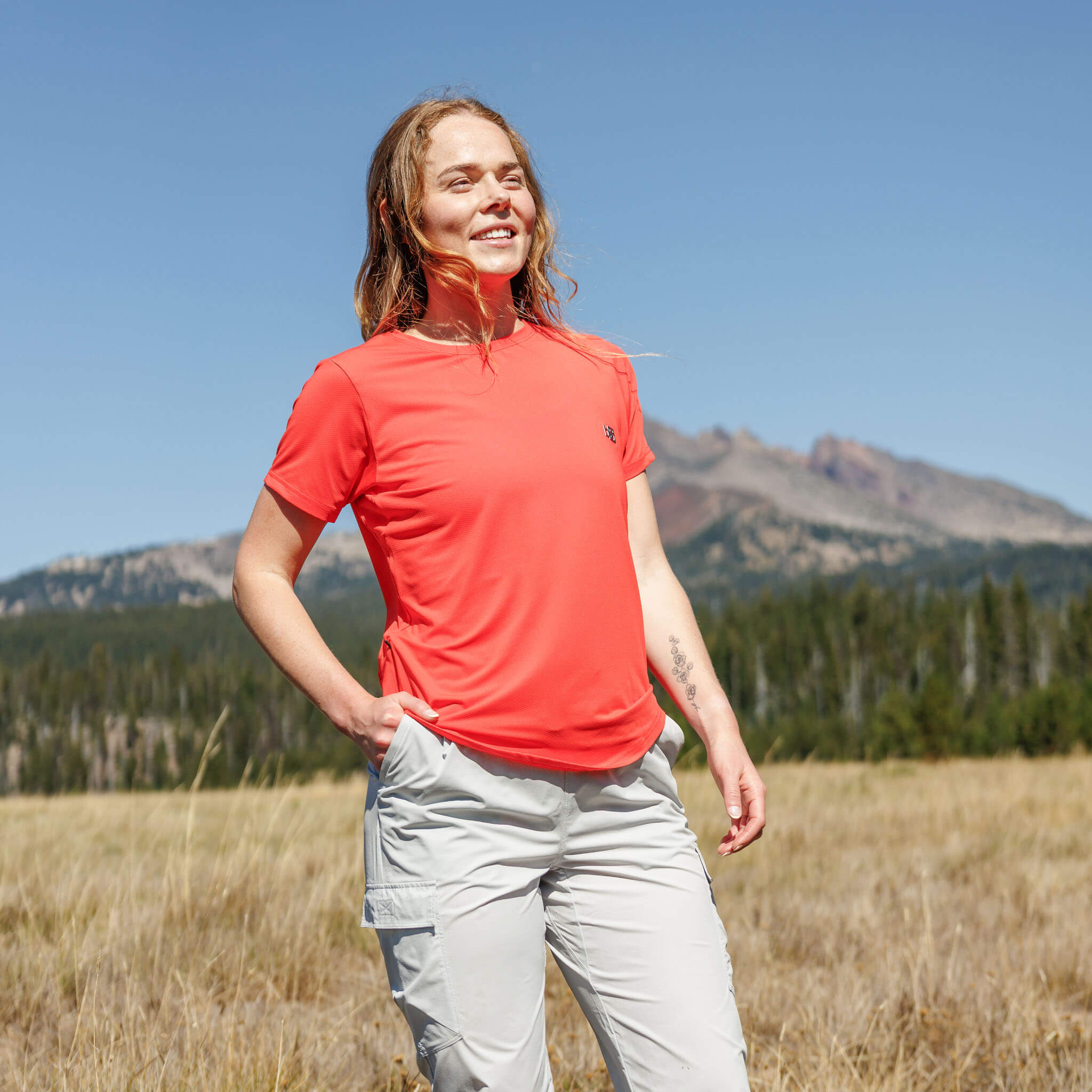 Woman standing in an open meadow with a mountain peak in the background wearing the BlackStrap Women's Voyage Tee