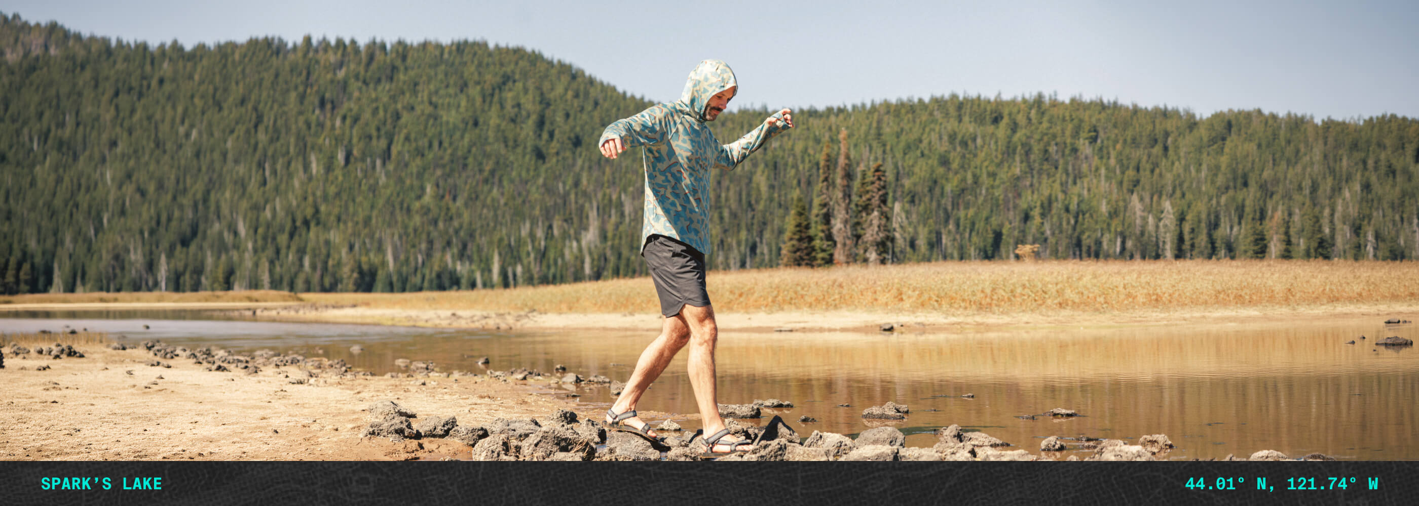 Man walking the shoreline wearing the BlackStrap Brackish Sun Hoodie and Evap Shorts as part of the Shoreline collection with a lake and trees in the background.