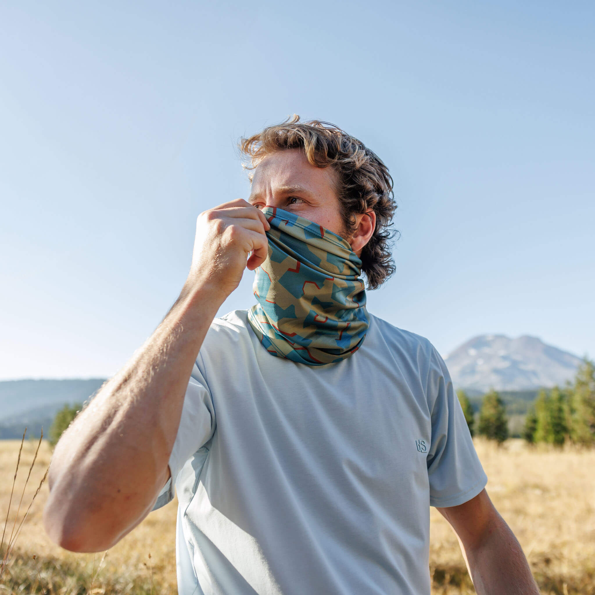 Man adjusting a BlackStrap Daily Tube neck gaiter over his nose with a mountain in the background #color_dimentical sagebrush