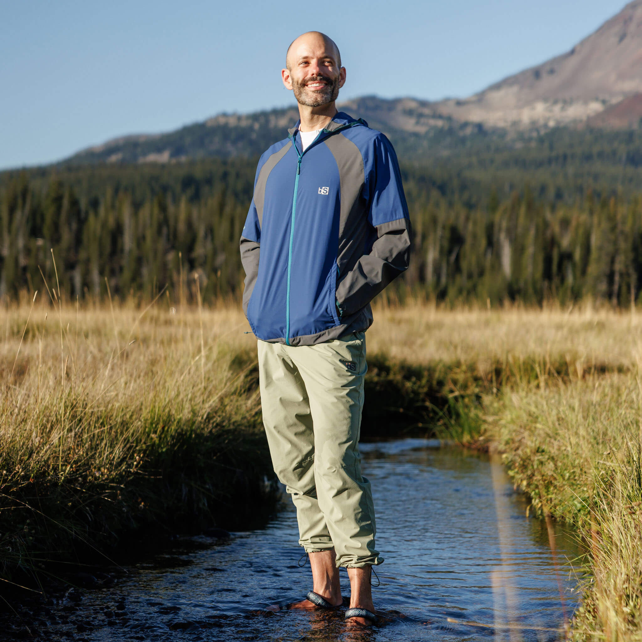Man smiling while standing in a shallow stream wearing BlackStrap approach pants with a Full Zip Cirq Jacket jacket, with a mountain meadow and peak visible in the background. #color_sagebrush
