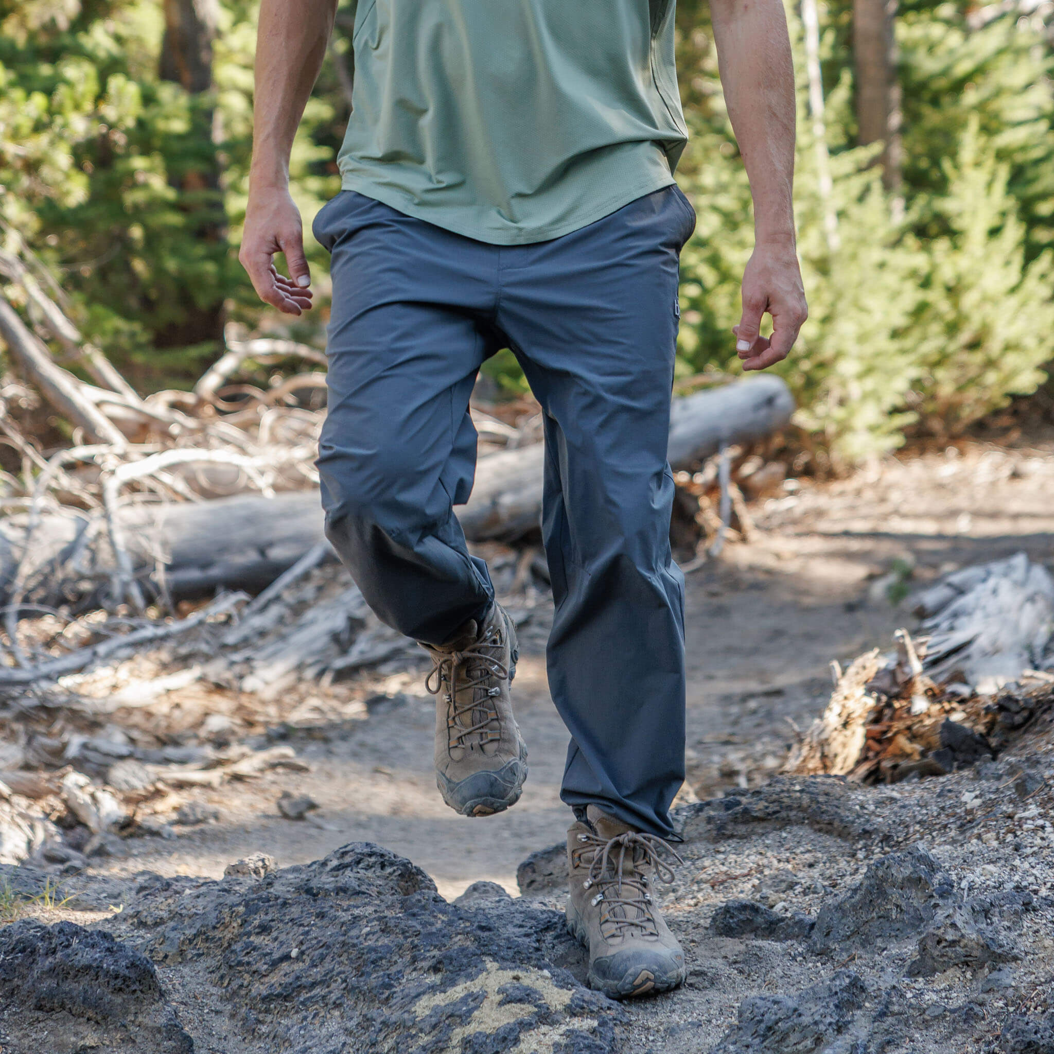 Man stepping over rocks on a forest trail wearing BlackStrap approach pants and hiking boots, paired with a Spruce Voyage short-sleeve shirt, with fallen logs and pine trees behind him. #color_smoldered