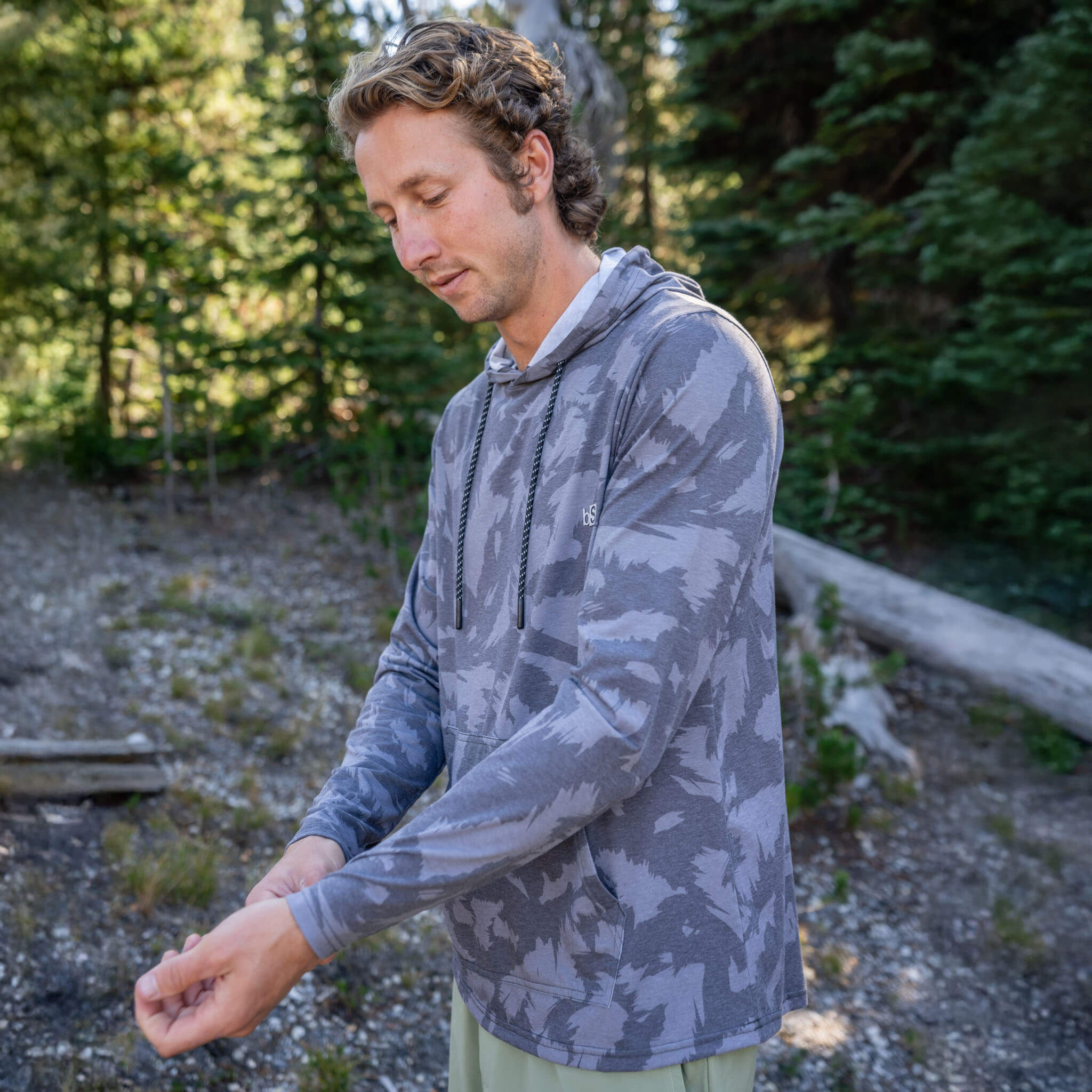 Man standing on a rocky trail in a pine forest wearing the BlackStrap Men's Aspect Hoodie Long Sleeve #color_slash smoldered