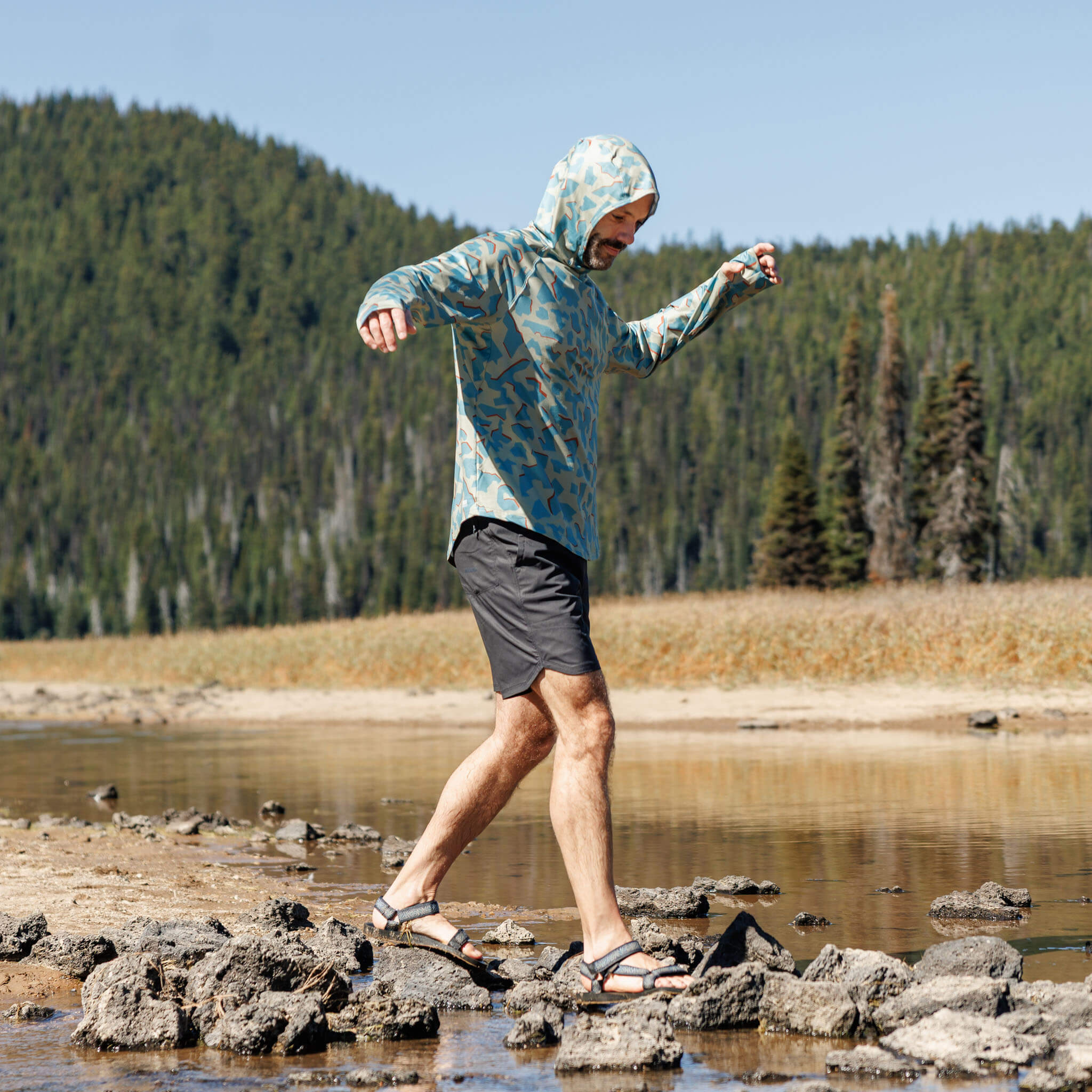 Man stepping across rocks along a lake's edge wearing the BlackStrap Men's Brackish Long Sleeve Hoodie with hood up #color_dimentical sagebrush