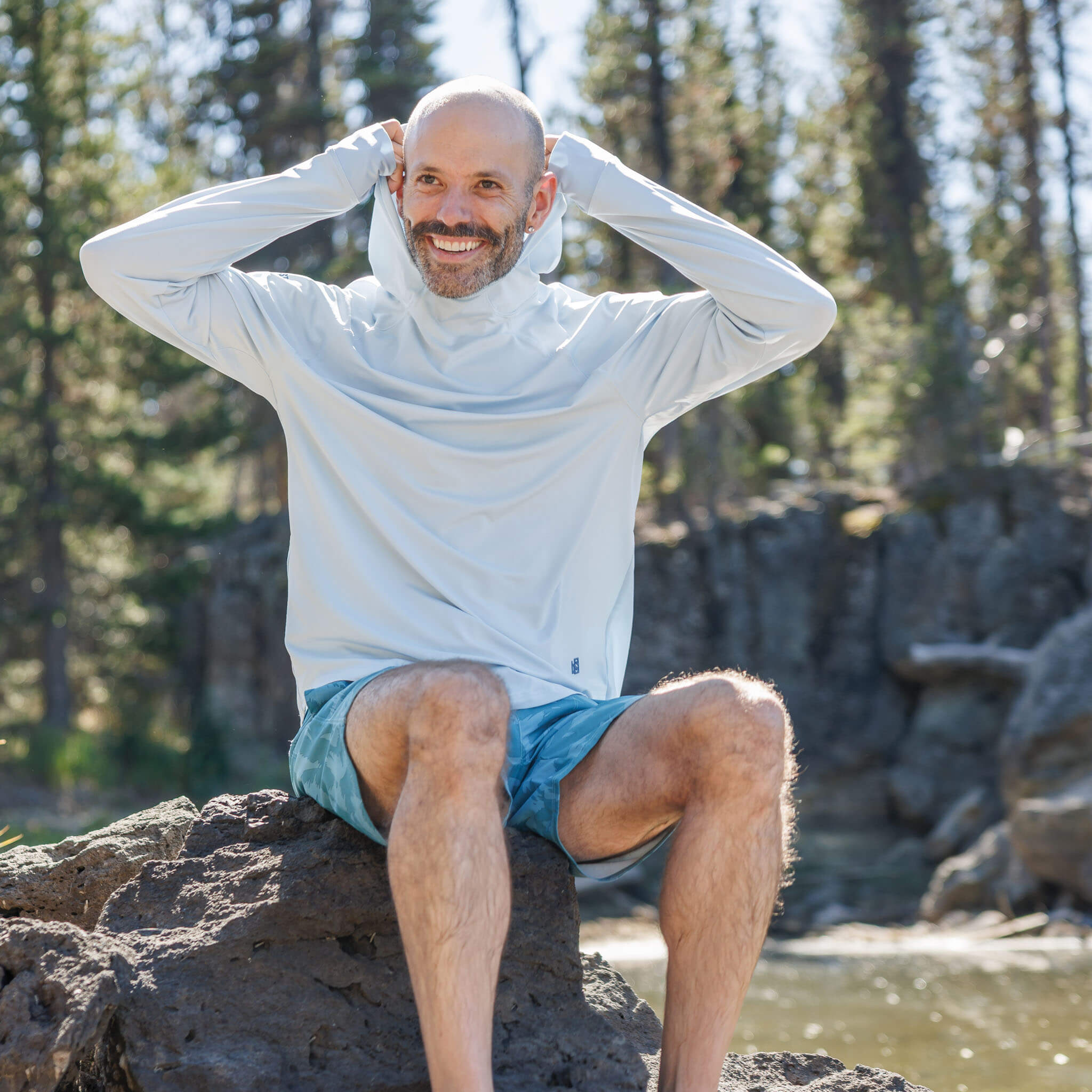 Man sitting on a rock by a mountain stream pulling up the hood of the BlackStrap Men's Brackish Long Sleeve Hoodie #color_stratus