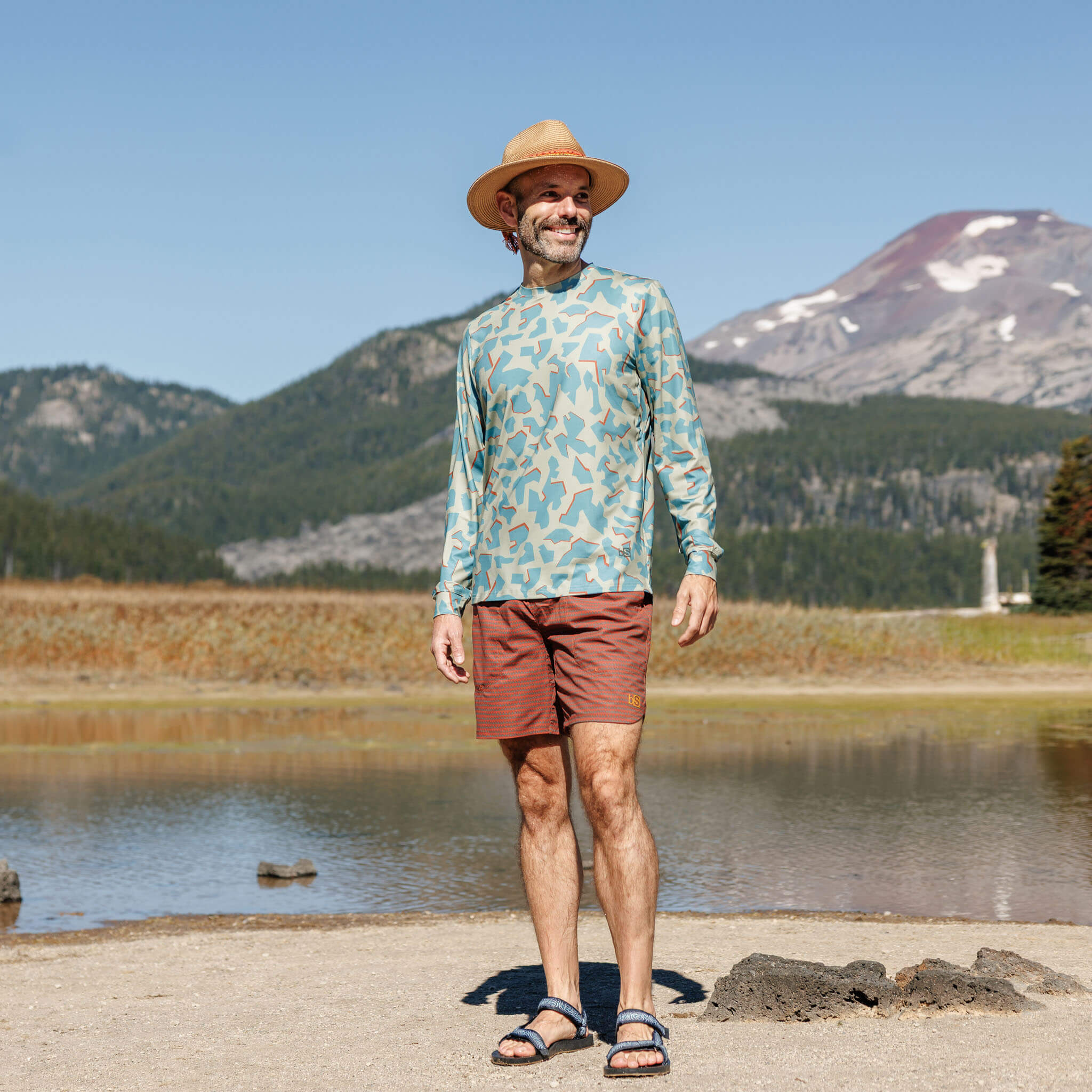 Man standing near an alpine lake with a snow-capped mountain in the background wearing the BlackStrap Men's Brackish Long Sleeve shirt