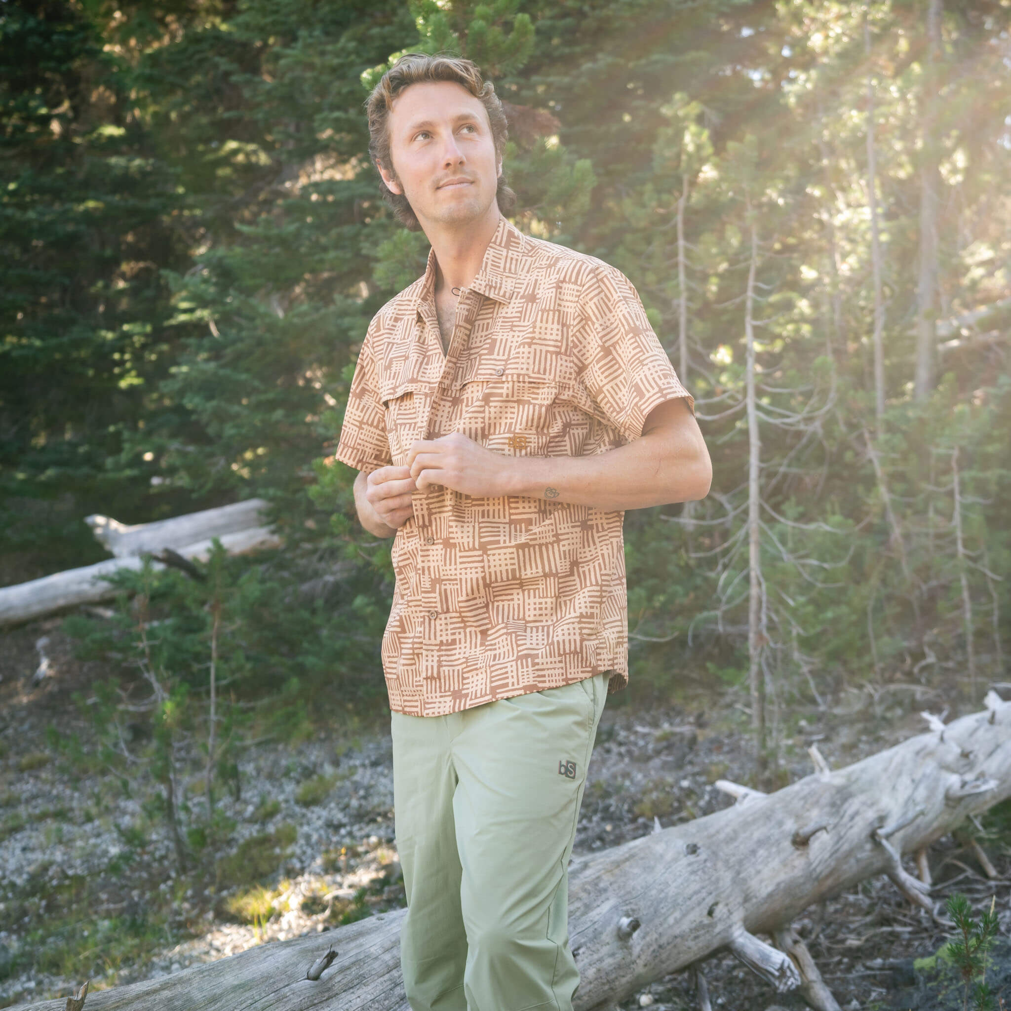 Man standing among pine trees buttoning a Scribble Lark short-sleeve BlackStrap camp shirt, paired with utility pants, with sunlight filtering through the forest behind him.
