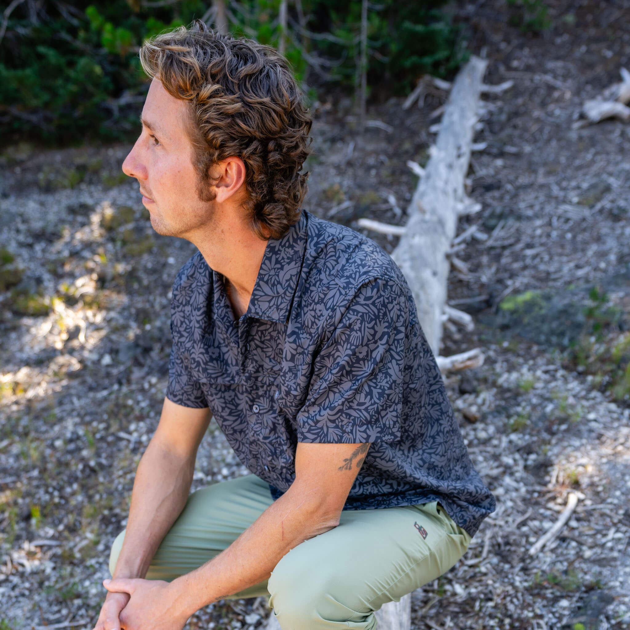 Man sitting outdoors in profile wearing a florican deschutes short-sleeve BlackStrap camp shirt and approach pants, with a rocky forest floor and fallen trees behind him