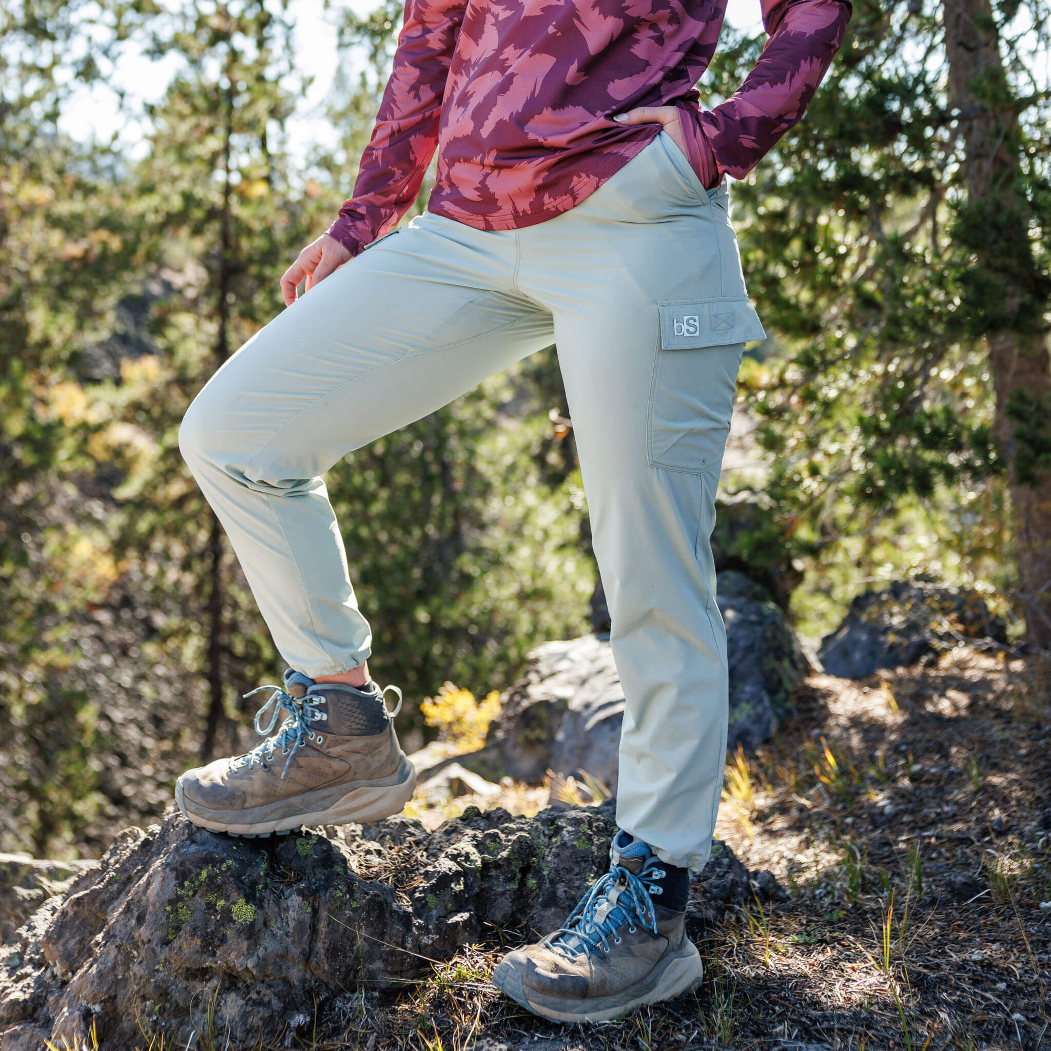 Woman posing on a boulder in a pine forest wearing BlackStrap approach pants with a cargo pocket and hiking boots, paired with a Voyage long-sleeve top #color_spruce