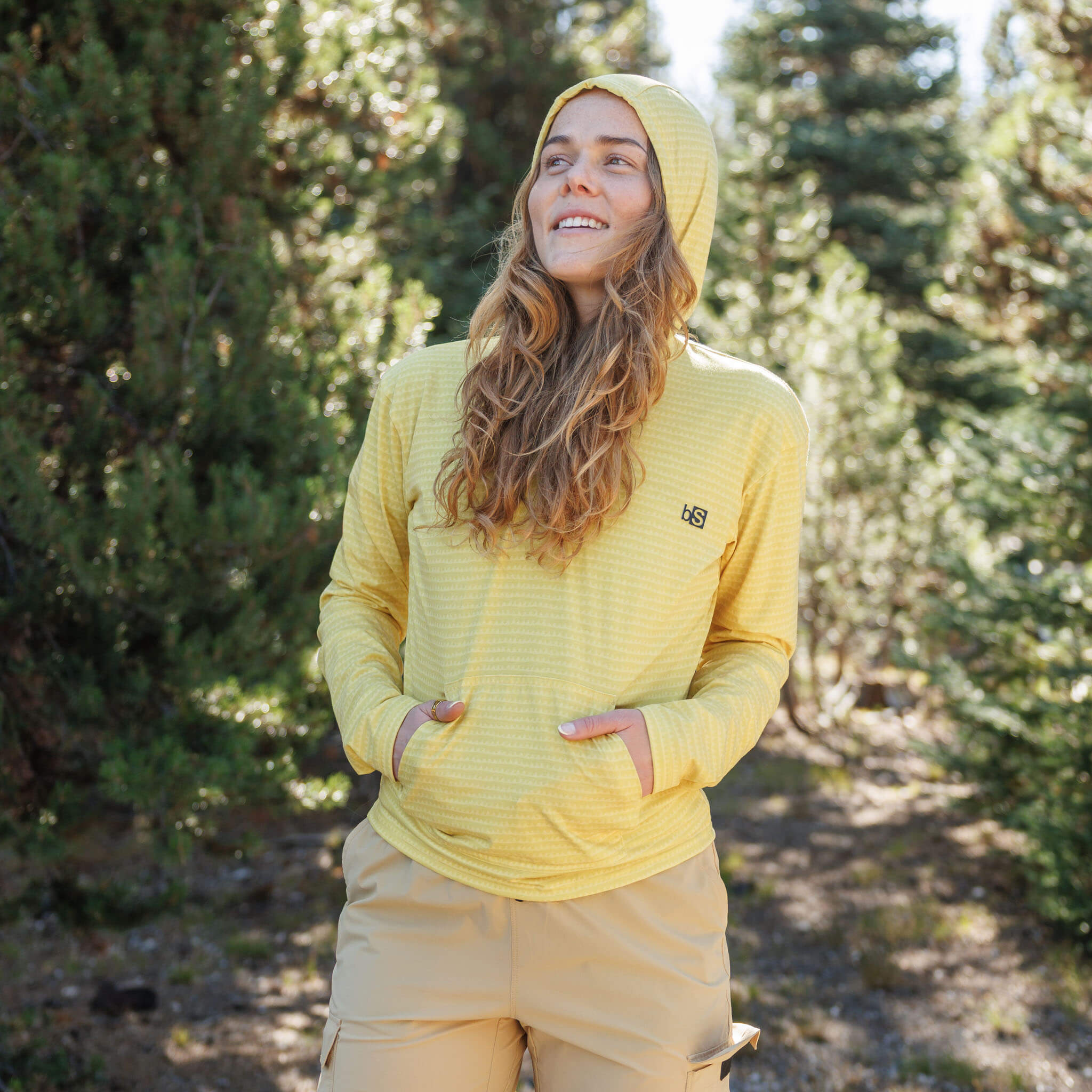 Woman standing on a pine forest trail with hands in the kangaroo pocket wearing the BlackStrap Women's Aspect Hoodie Long Sleeve #color_sharktooth meadow