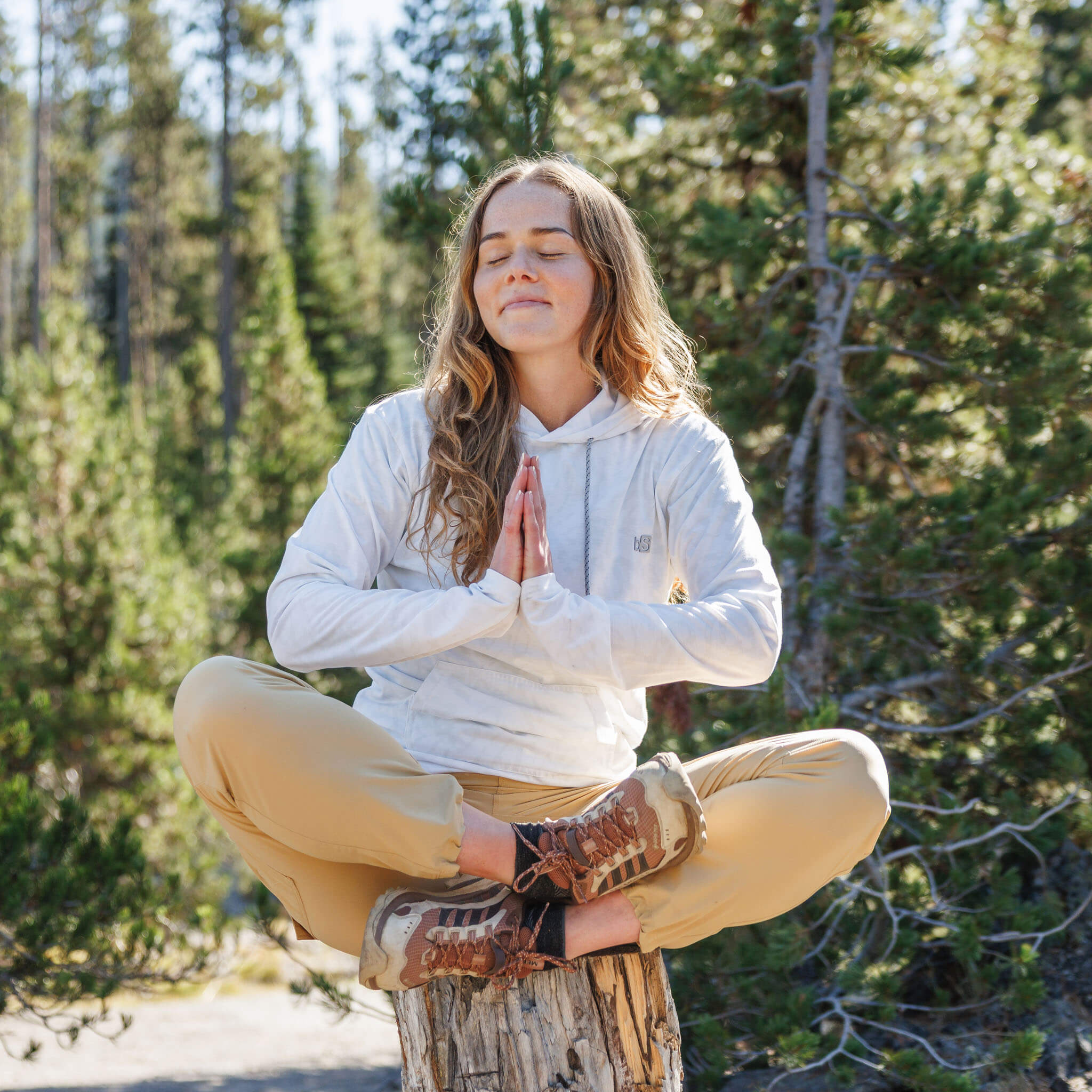 Woman meditating on a tree stump in a pine forest wearing the BlackStrap Women's Aspect Hoodie Long Sleeve