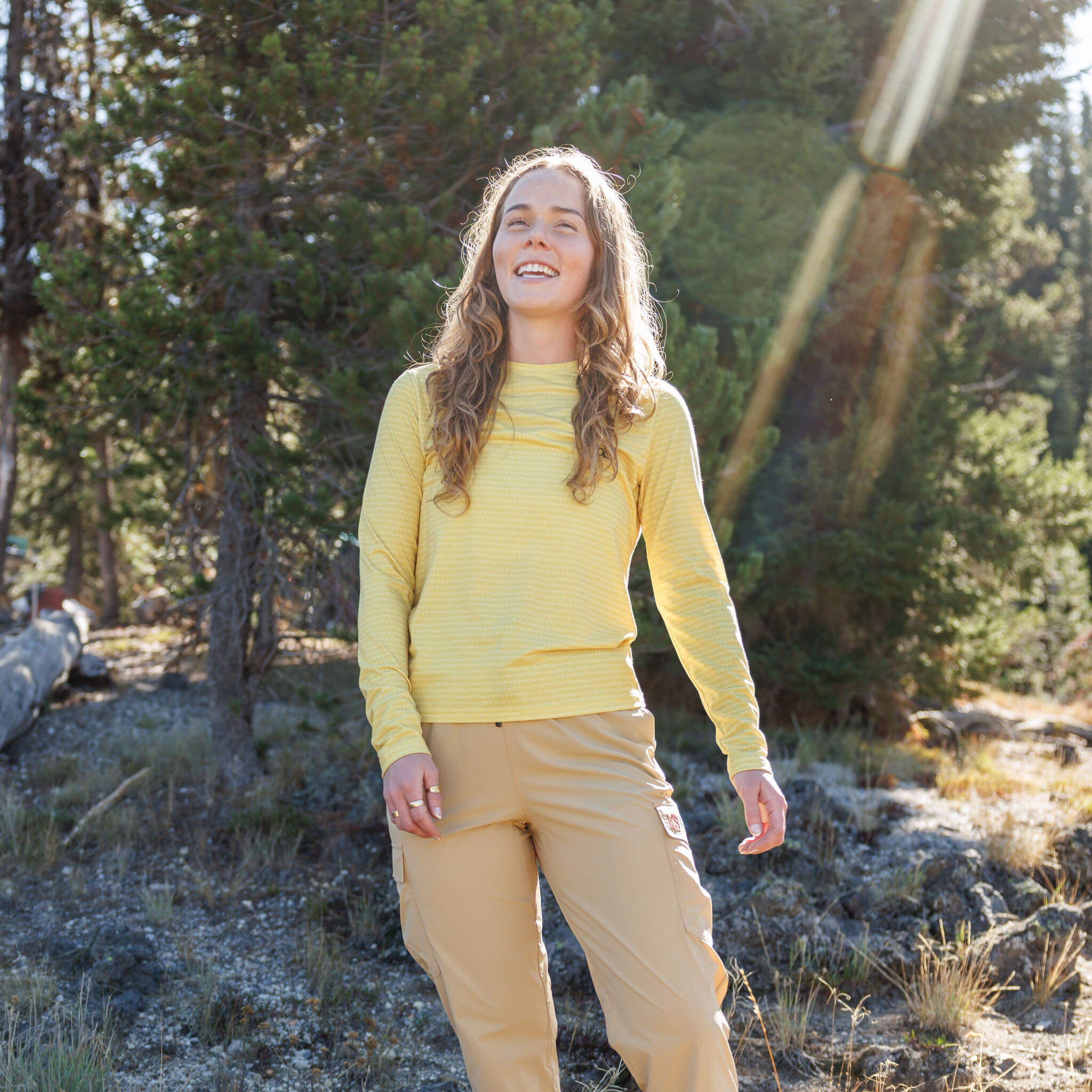 Woman smiling in a sunlit alpine clearing wearing the BlackStrap Women's Aspect Long Sleeve #color_sharktooth meadow