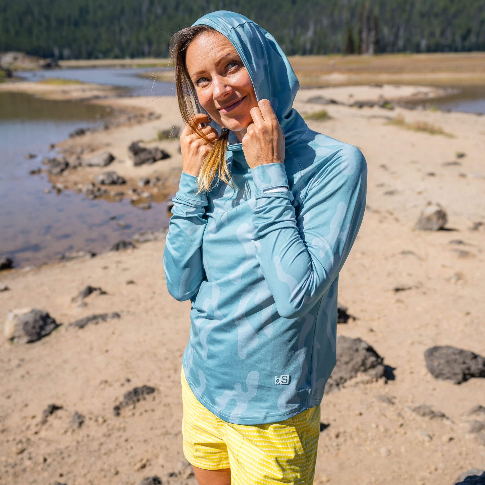 Women standing on a sandy riverbank adjusting the hood of the BlackStrap Women's Brackish Long Sleeve Hoodie #color_corally surge