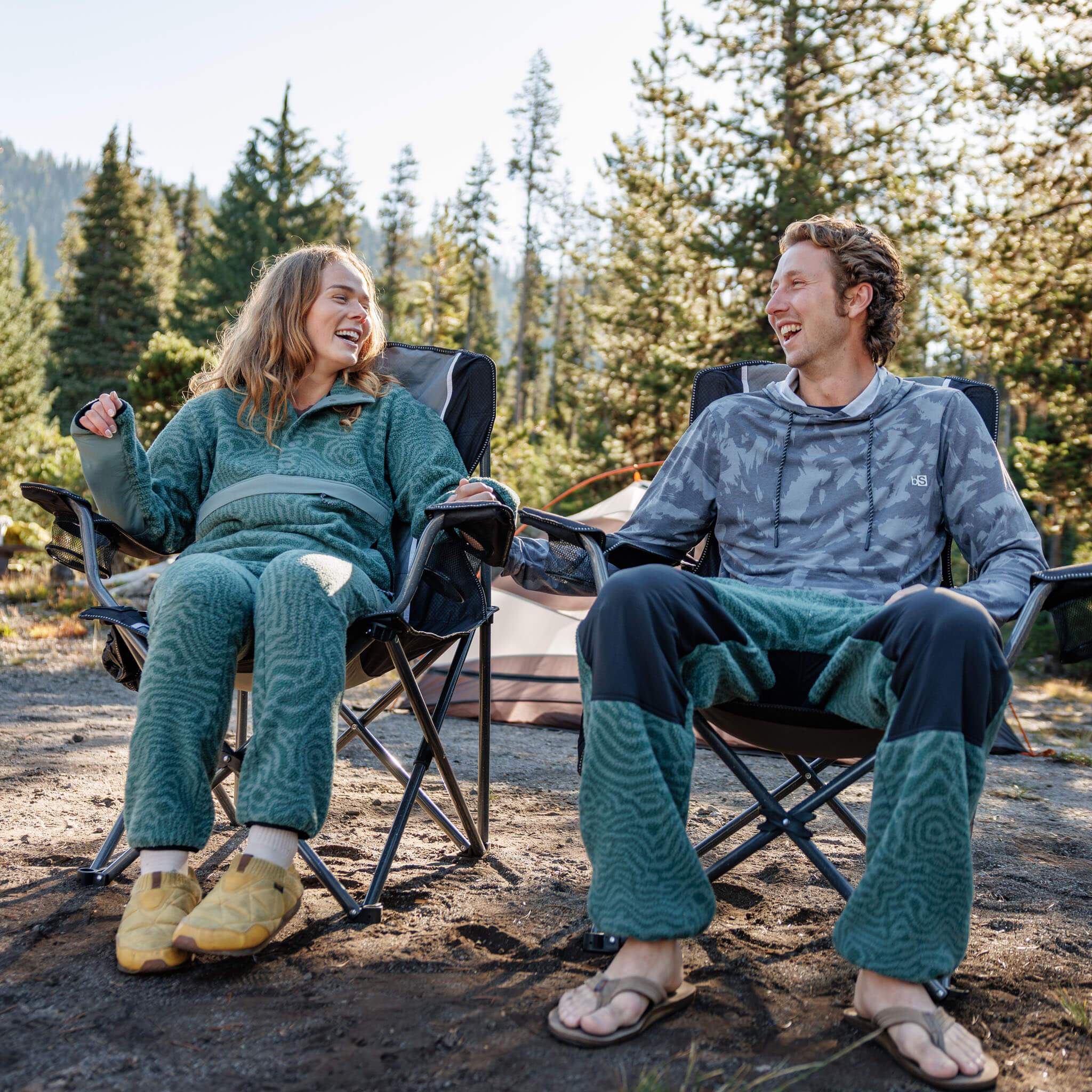 A woman in the BlackStrap Women's Caldera Sherpa Fleece set and a man in the BlackStrap Men's Aspect Hoodie relaxing in camp chairs at a pine forest campsite
