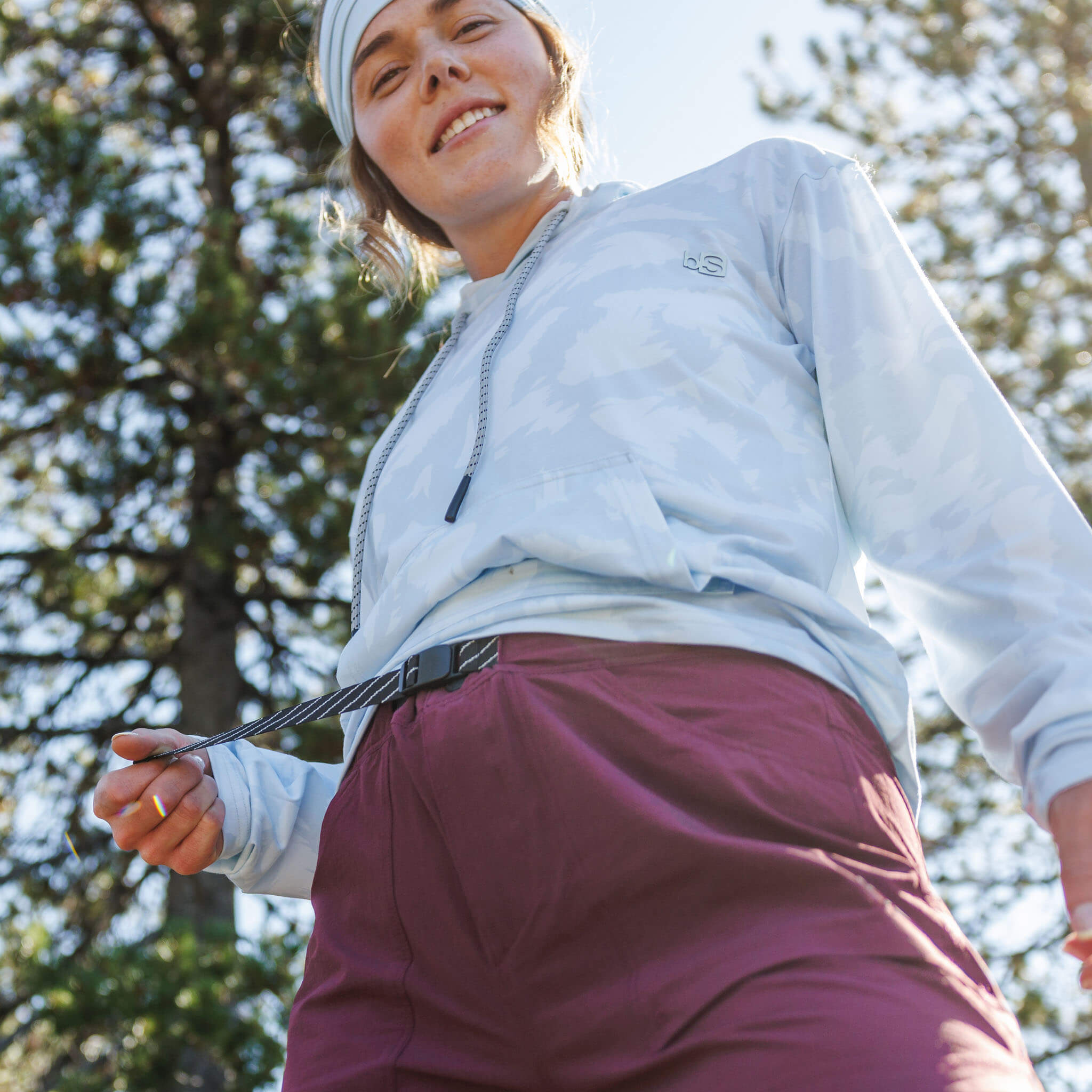 Woman smiling outdoors holding the end of a striped buckle belt on utility shorts, wearing a BlackStrap Aspect hoodie and Multifunctional headband, with pine trees behind her. #color_cherry