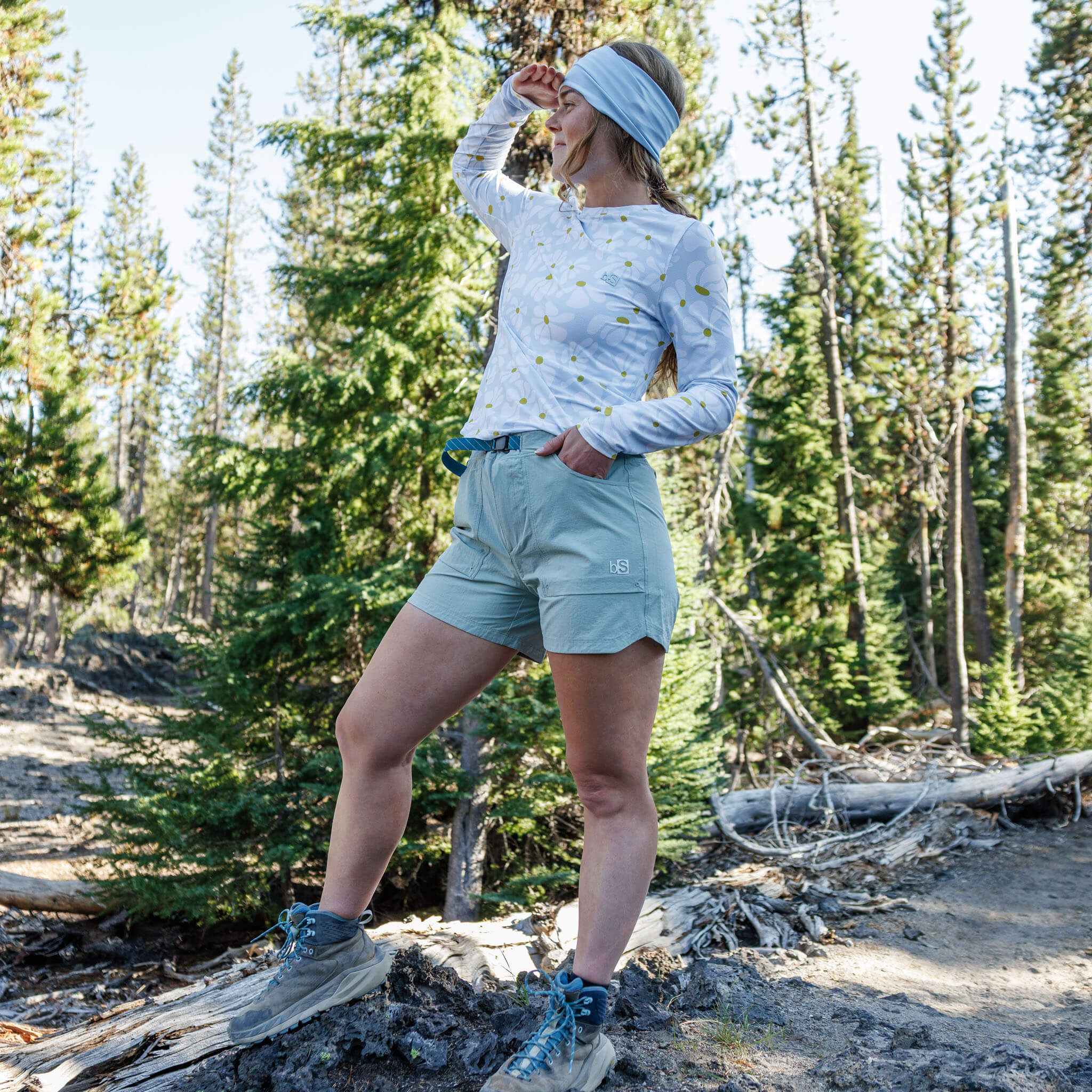 Woman standing on a log in a pine forest wearing utility shorts with a belt, a long-sleeve voyage crewneck trail shirt, a multifunctional headband, and hiking boots.