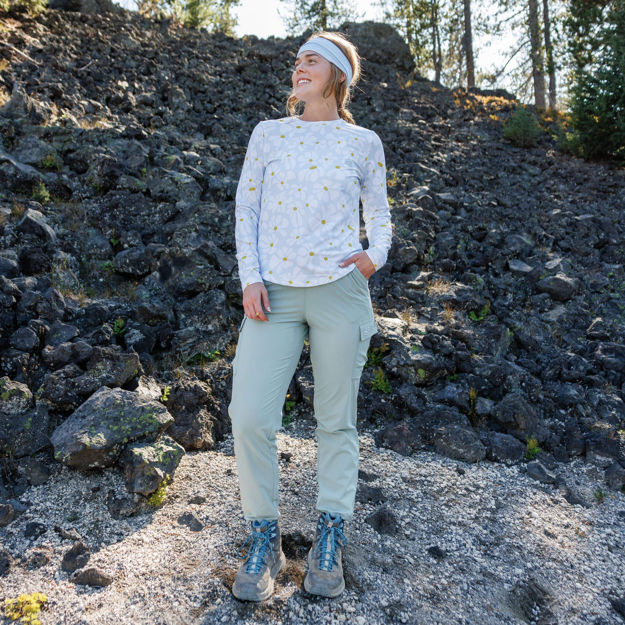 Woman standing on a volcanic rock hillside wearing the BlackStrap Women's Voyage Crew Long Sleeve #color_boho stratus